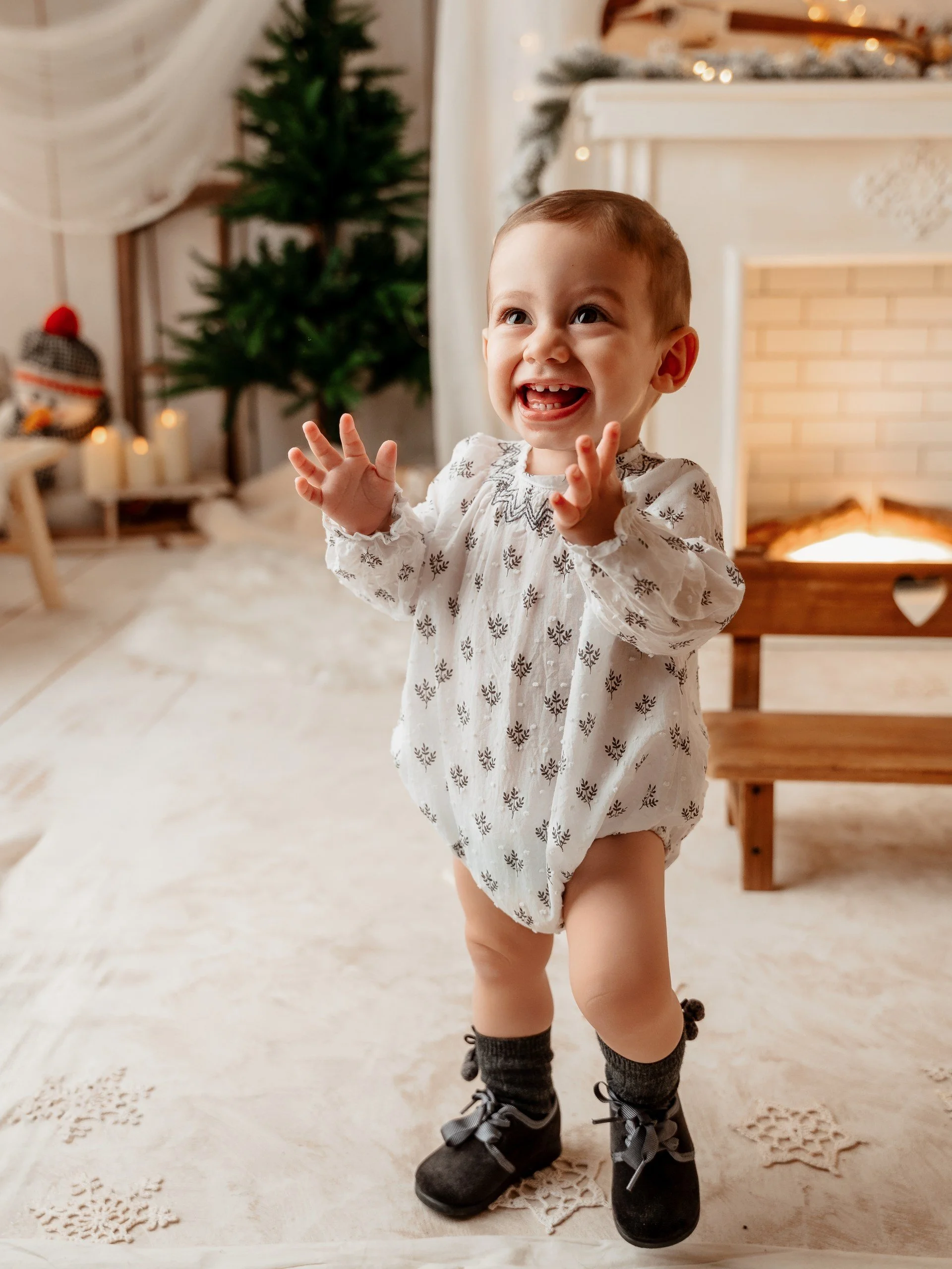 Niño pequeño con ropa de pijama, botas negras, en un ambiente decorado para Navidad con un árbol y una chimenea