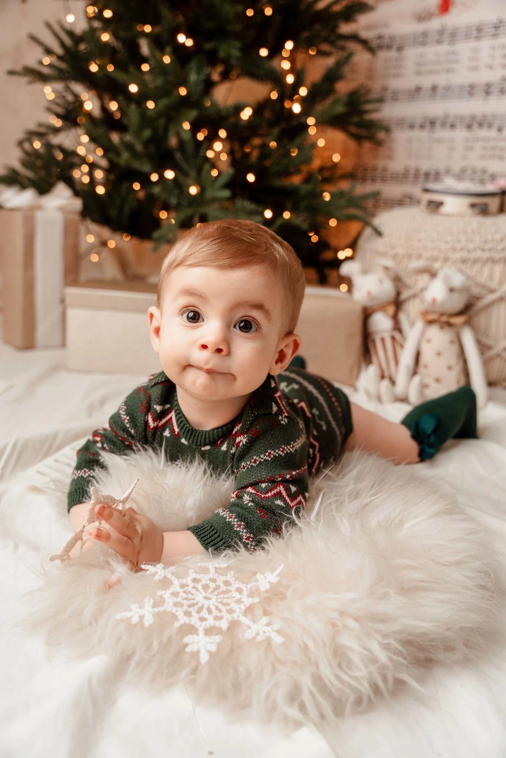 Un niño con ropa navideña acostado en una manta blanca con estrella de encaje, en un ambiente decorado con un árbol de Navidad con luces, regalos y figuras navideñas.