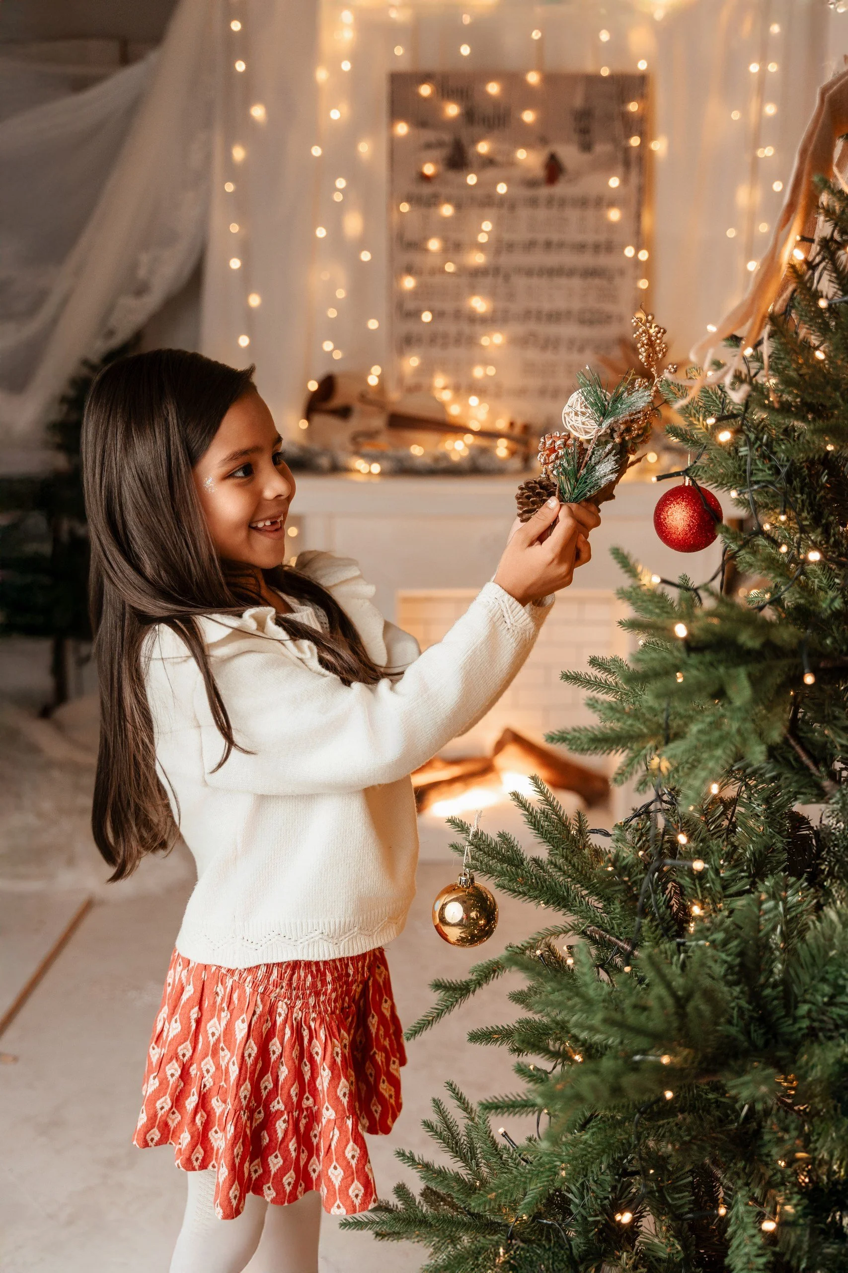 Niña con vestido rojo y suéter blanco decorando un árbol de Navidad con esferas doradas y adornos. En el fondo hay luces decorativas y un cartel con notas musicales.