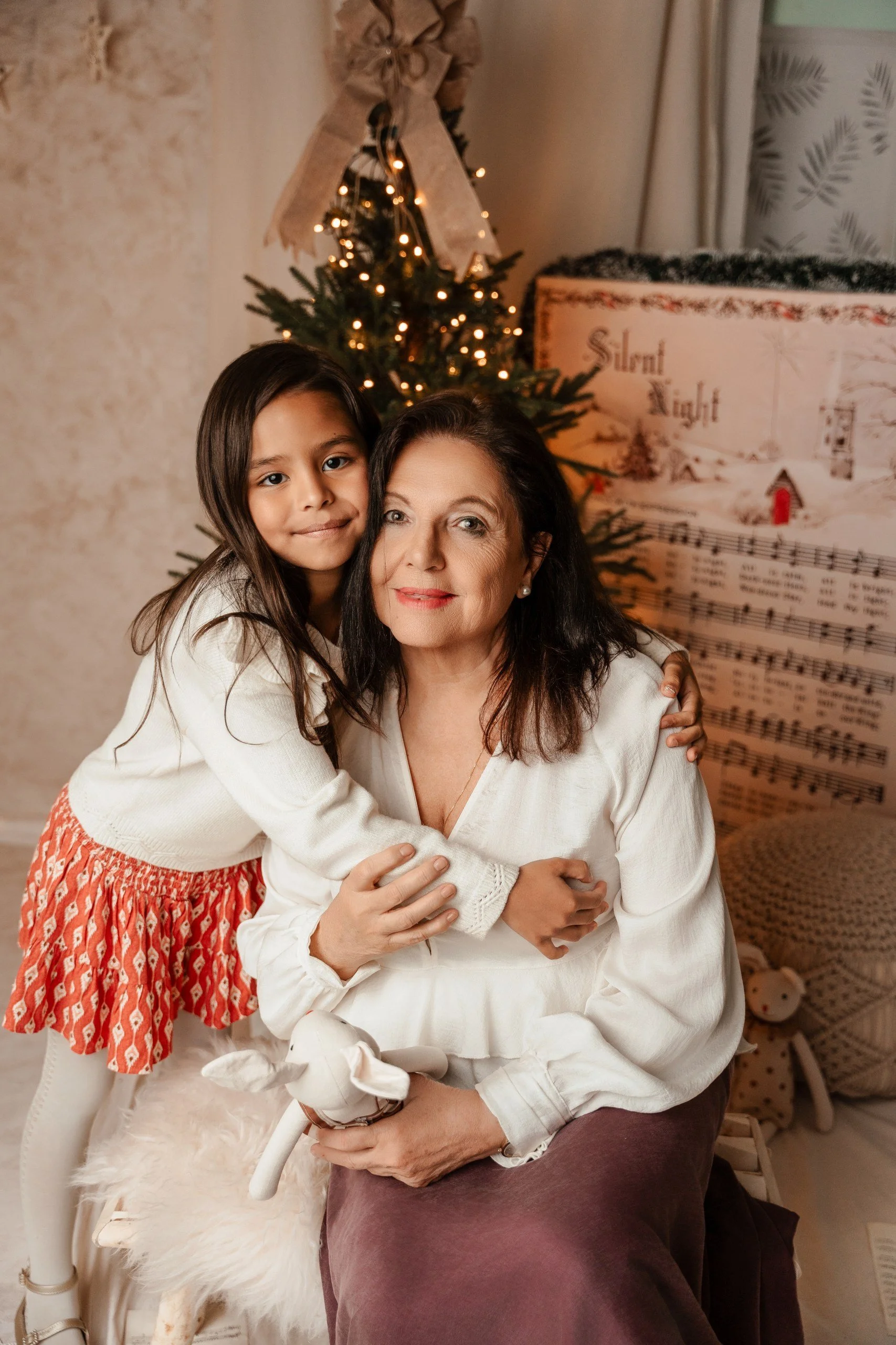 Una niña y una mujer en un ambiente navideño, con un árbol decorado con luces y un lazo grande en la parte superior. La mujer está sentada en una silla y la niña la abraza con cariño. Hay una decoración temática con música y un muñeco de nieve en el 