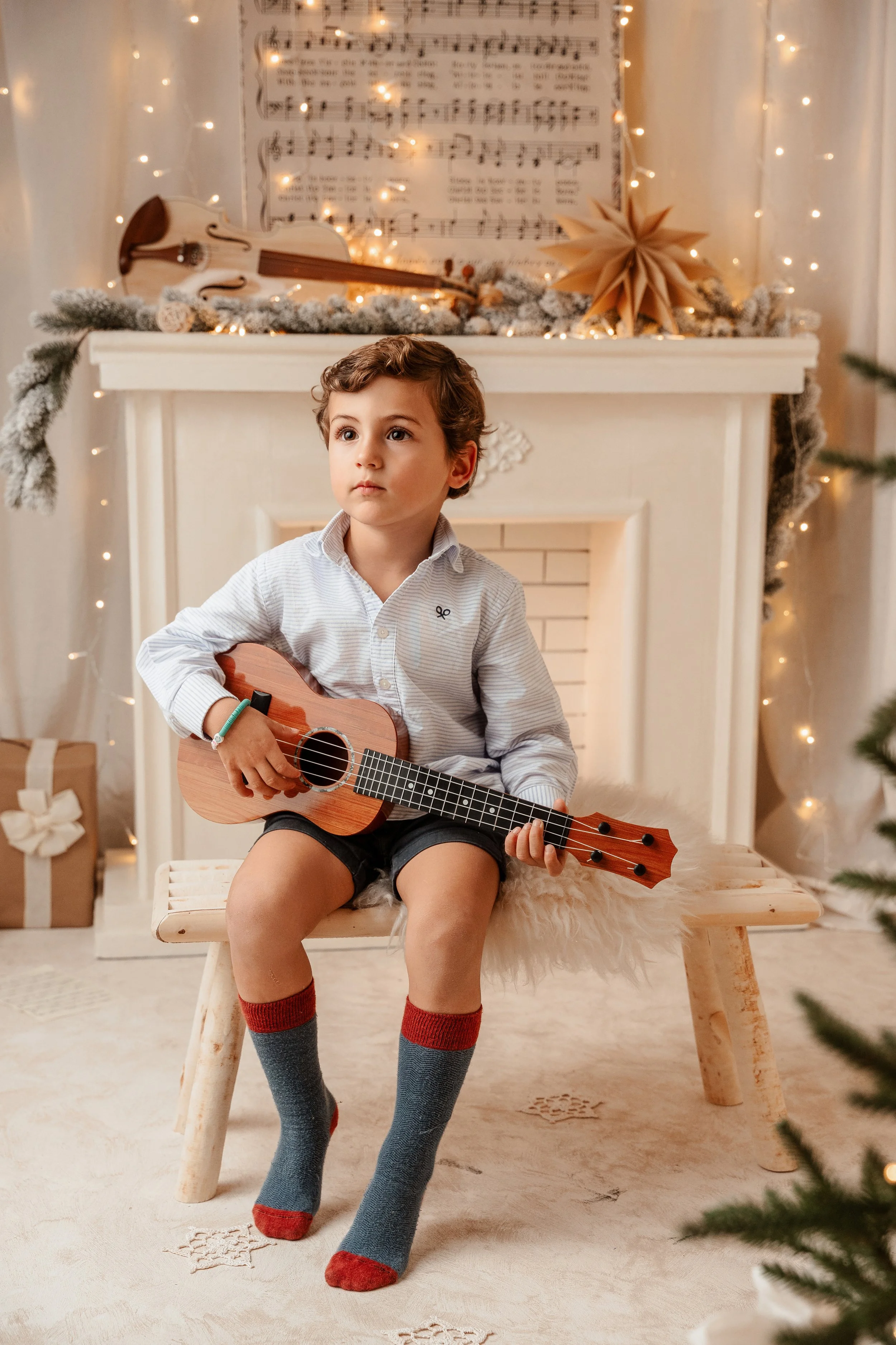 Un niño sentado en un banco de madera, tocando una guitarra acústica, con decoración navideña en el fondo, incluyendo una gran estrella de papel, luces y regalos envueltos, en un ambiente festivo y cálido.