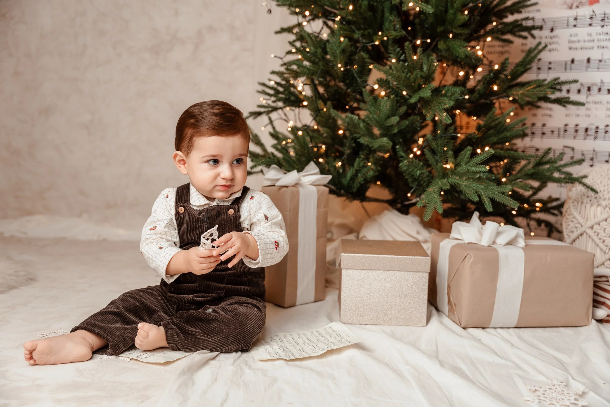 Un niño pequeño sentado en el suelo en una sala decorada con un árbol de Navidad iluminado y regalos envueltos, sosteniendo una pieza de encaje en las manos, mientras mira hacia un lado con expresión pensativa.
