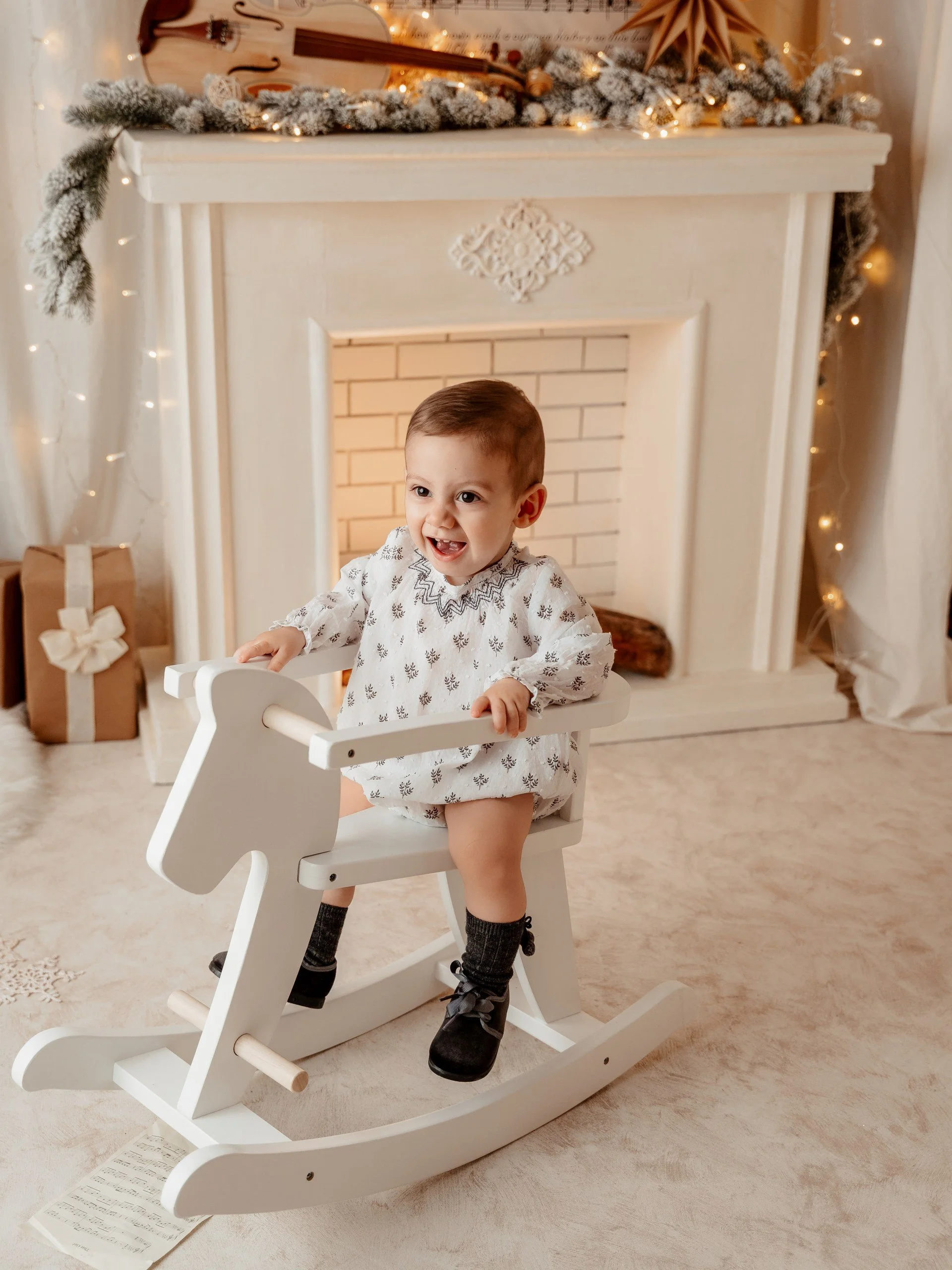 Un niño pequeño montado en un caballito de madera blanco en el interior de una sala decorada con motivos navideños, un chimenea, regalos y adornos