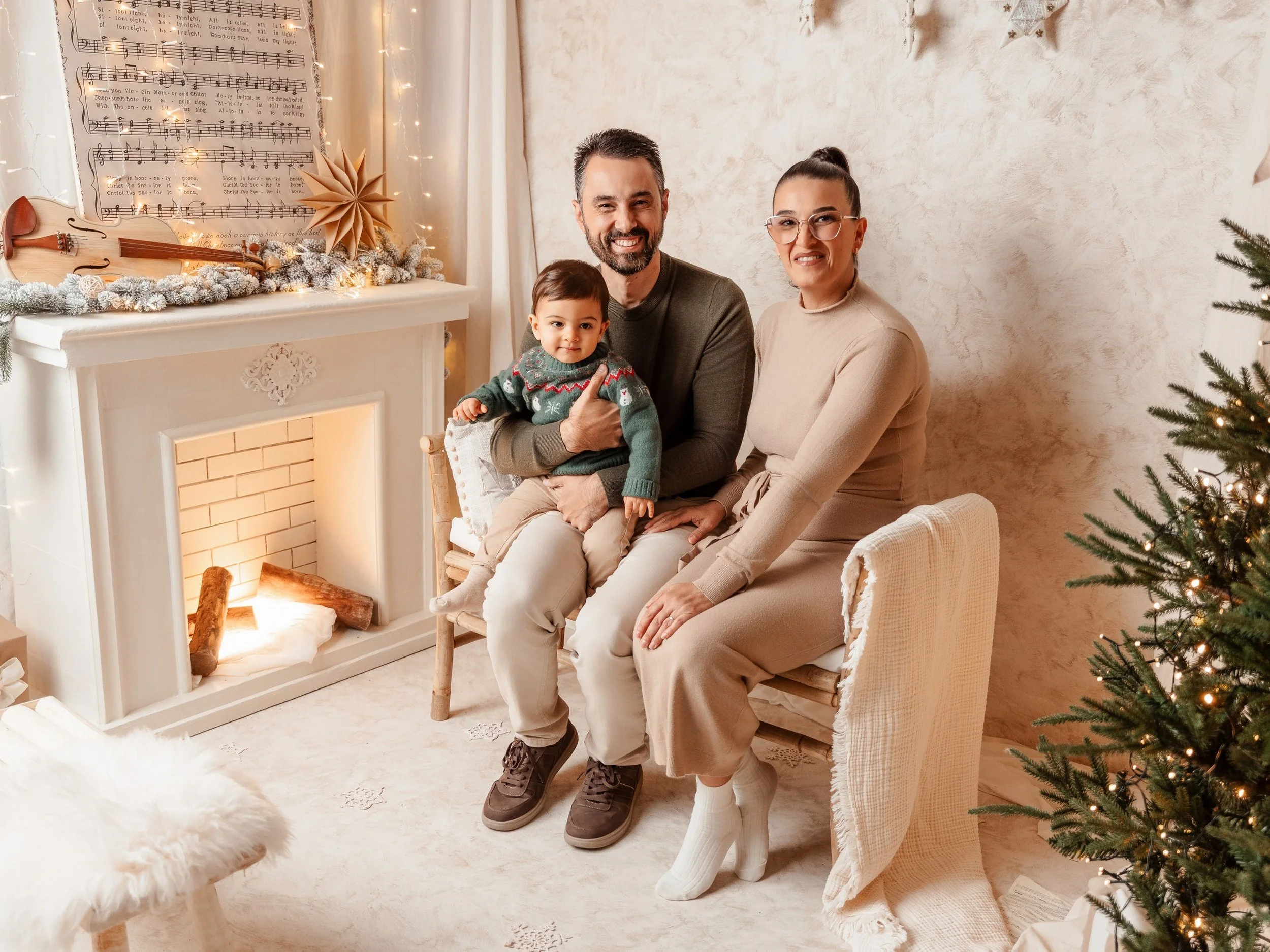 Una familia de tres personas, un hombre, una mujer y un niño pequeño, posando en una sala decorada para Navidad, con un árbol navideño y una chimenea decorativa.
