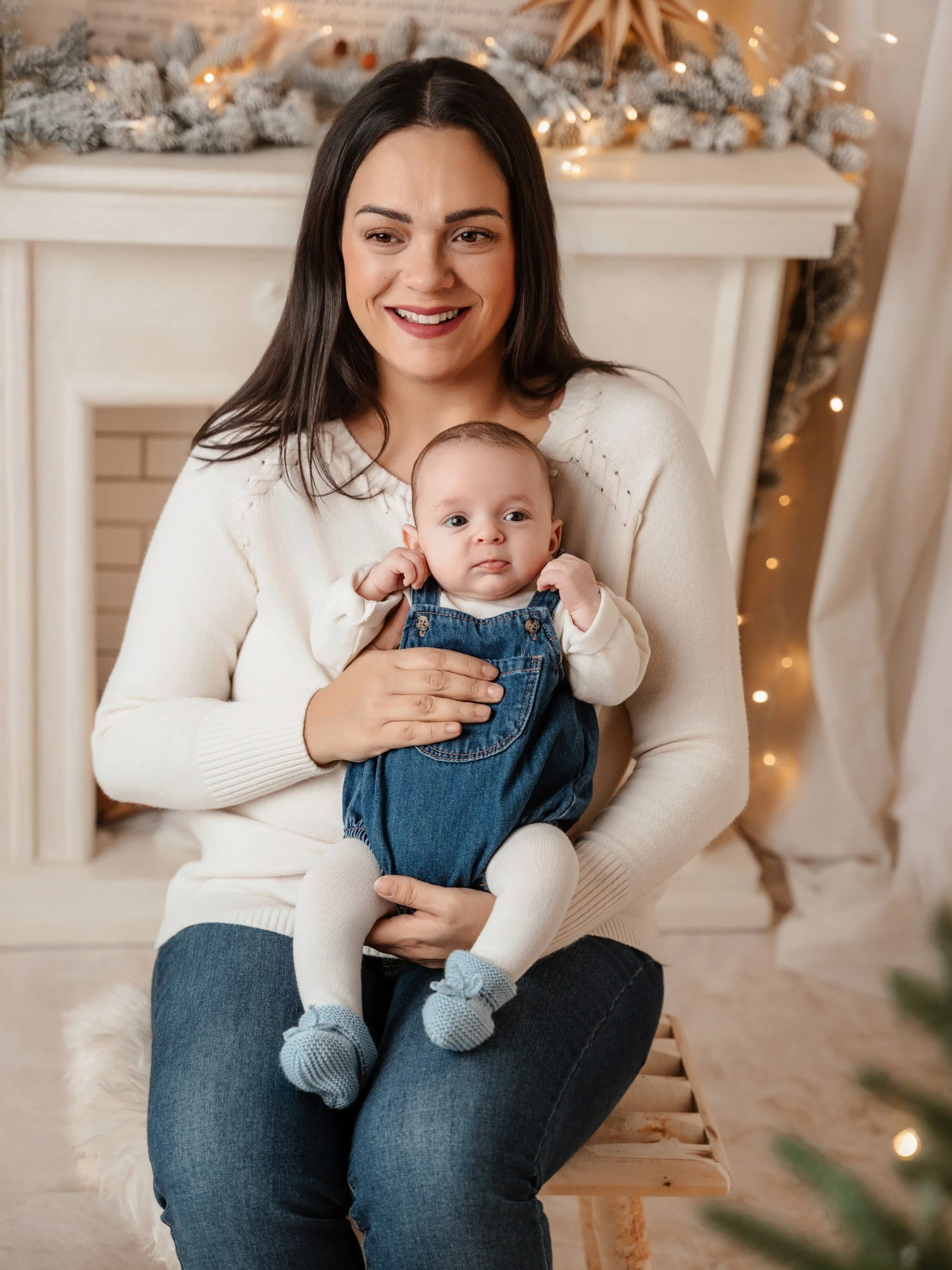 Mujer con cabello oscuro y blusa blanca, sosteniendo a un bebé con ropa azul, en una decoración navideña con árbol y luces.