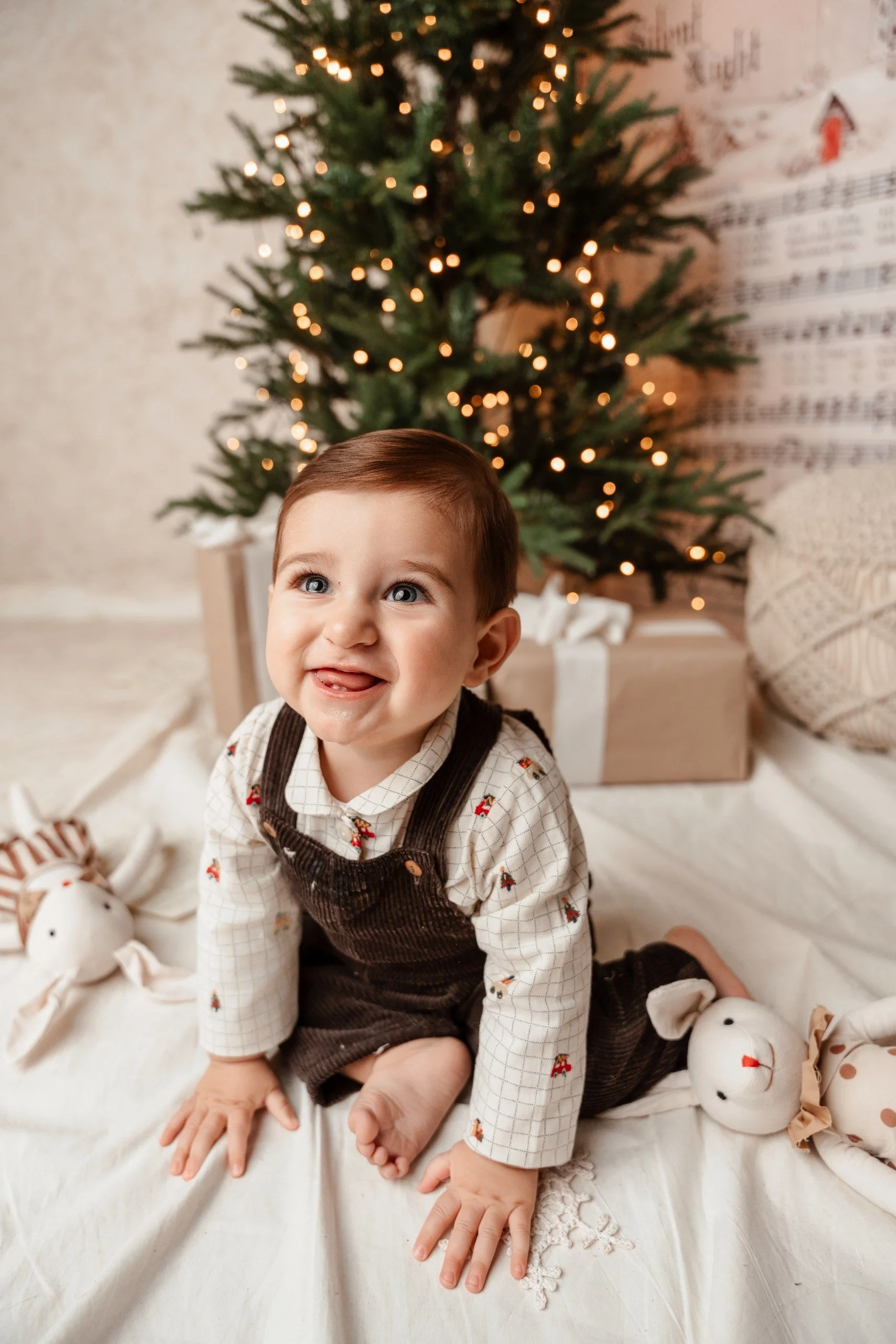 Un niño pequeño con cabello castaño, ojos azules, vistiendo una camisa de cuadros con detalles navideños y un overol café, en posición de gateo delante de un árbol de Navidad decorado con luces, con figuras de muñecos de nieve y regalos en el fondo.