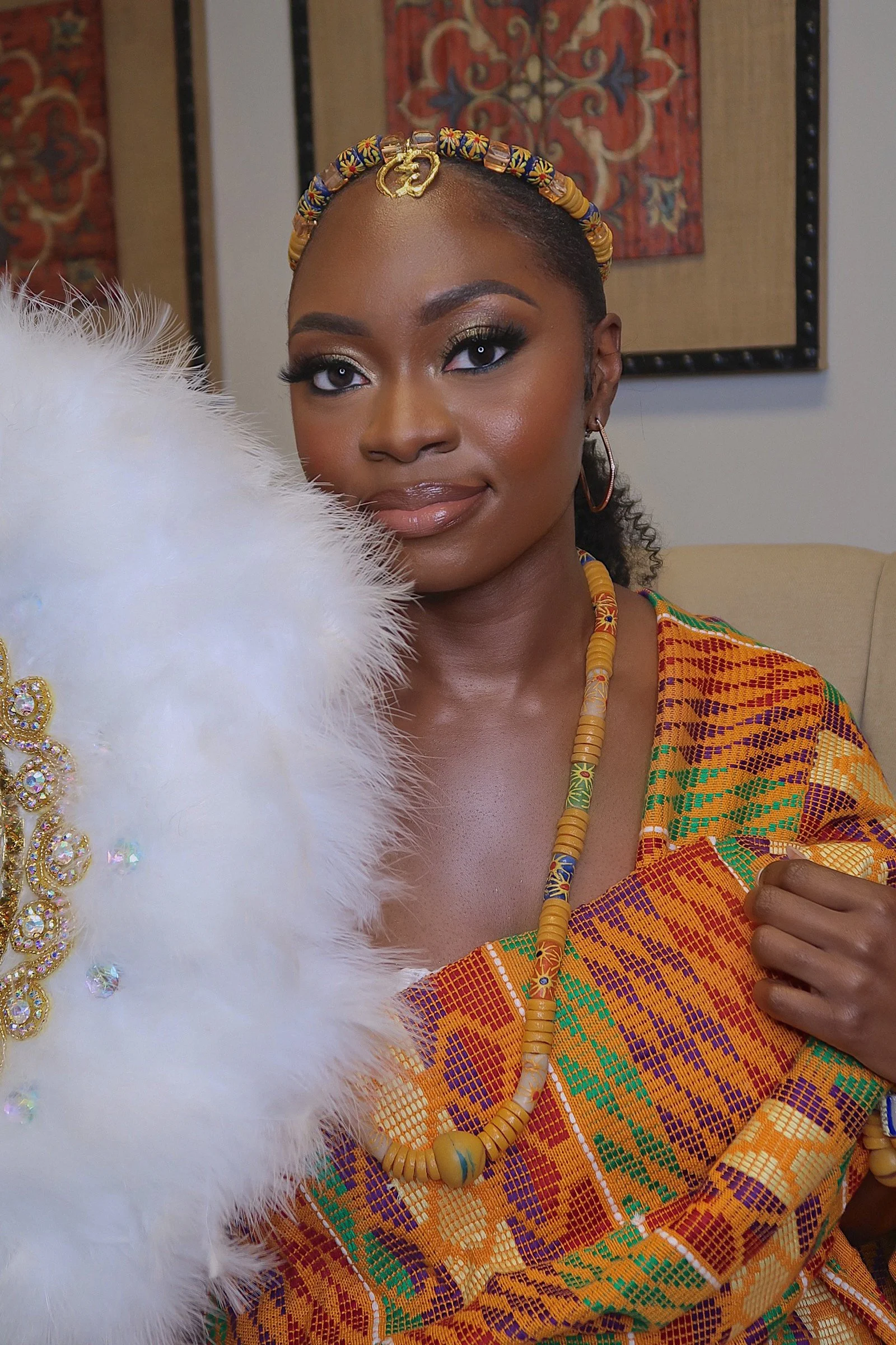 A woman dressed in traditional African attire with colorful beads and jewelry, holding a white feathered accessory, in a room with patterned wall art.
