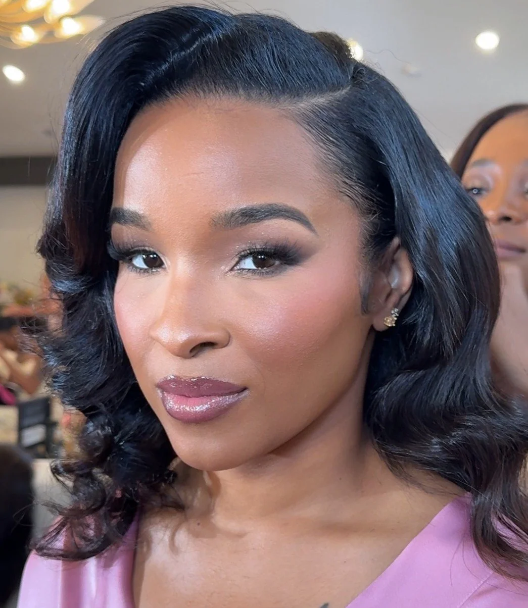 Close-up of a woman with long braids, glamorous makeup, large rhinestone hoop earrings, and a colorful patterned top. She is touching her head with her hand and looking slightly upward.