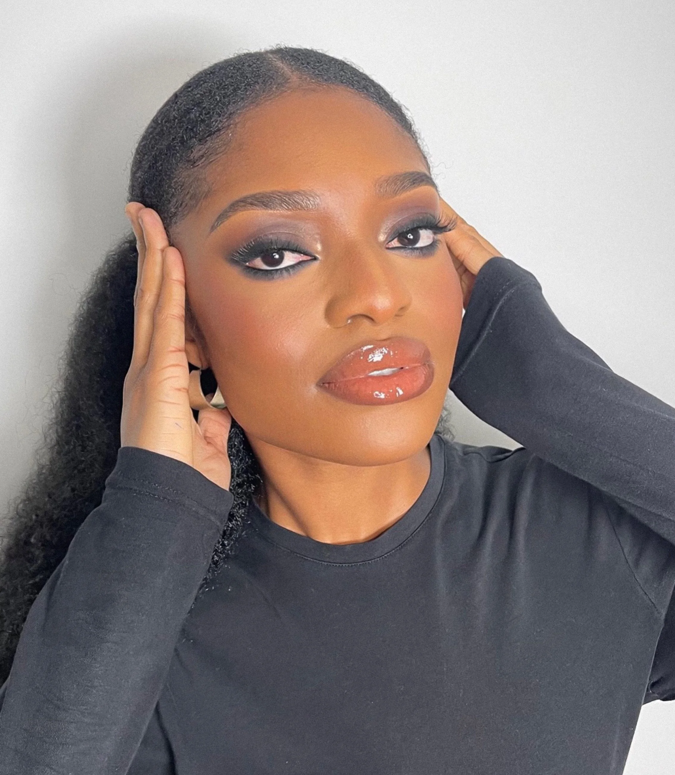 Close-up portrait of a woman with dark curly hair, wearing makeup with smoky eyeshadow and glossy lipstick, posing with her hands near her head, against a plain light background.