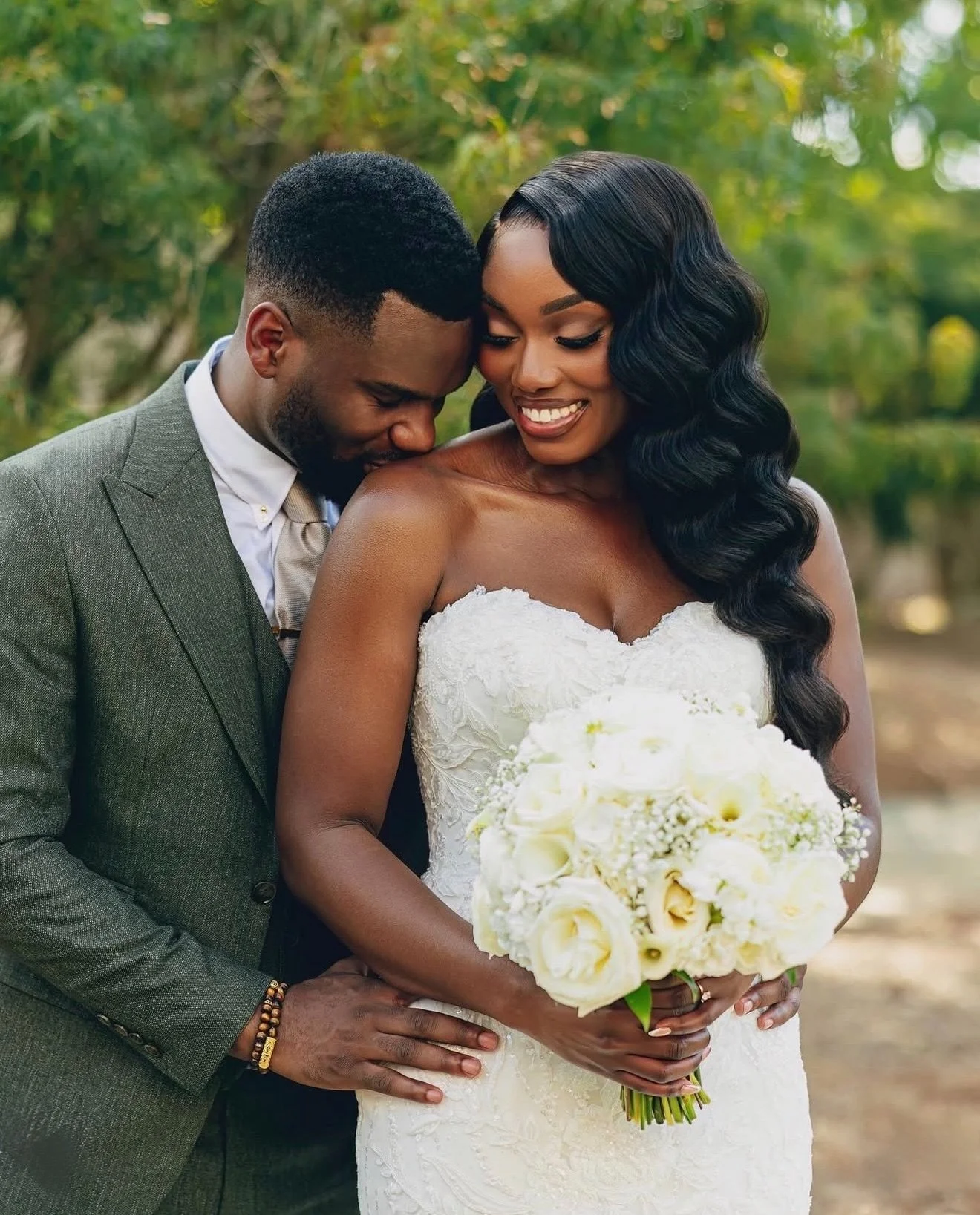 A woman with long, braided hair styled in twists, wearing makeup, gold earrings, and layered necklaces, posing indoors.