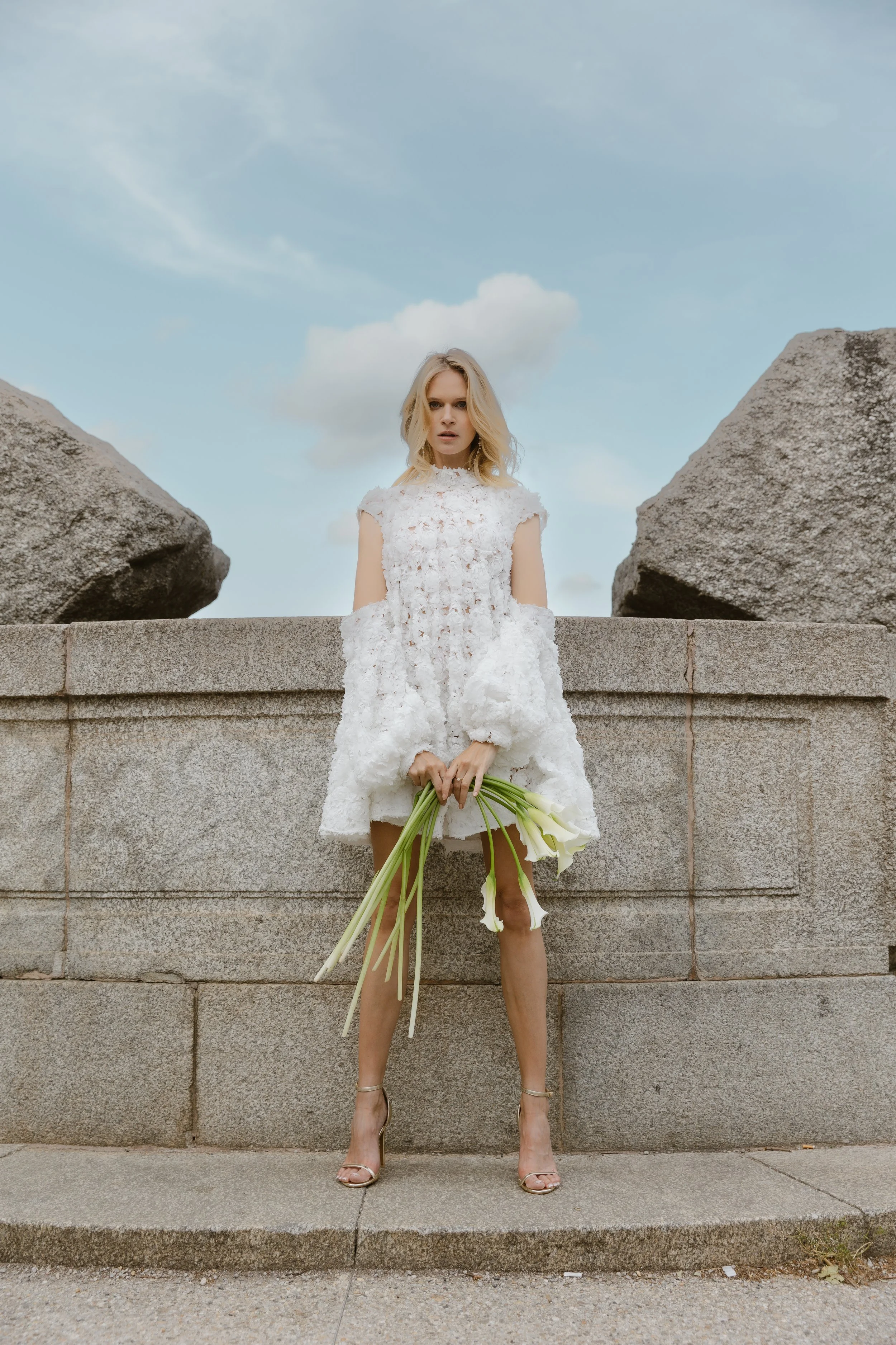 Frau in weißem Kleid hält Blumen vor einer Steinmauer mit großen Steinen im Hintergrund, blauer Himmel mit Wolken.