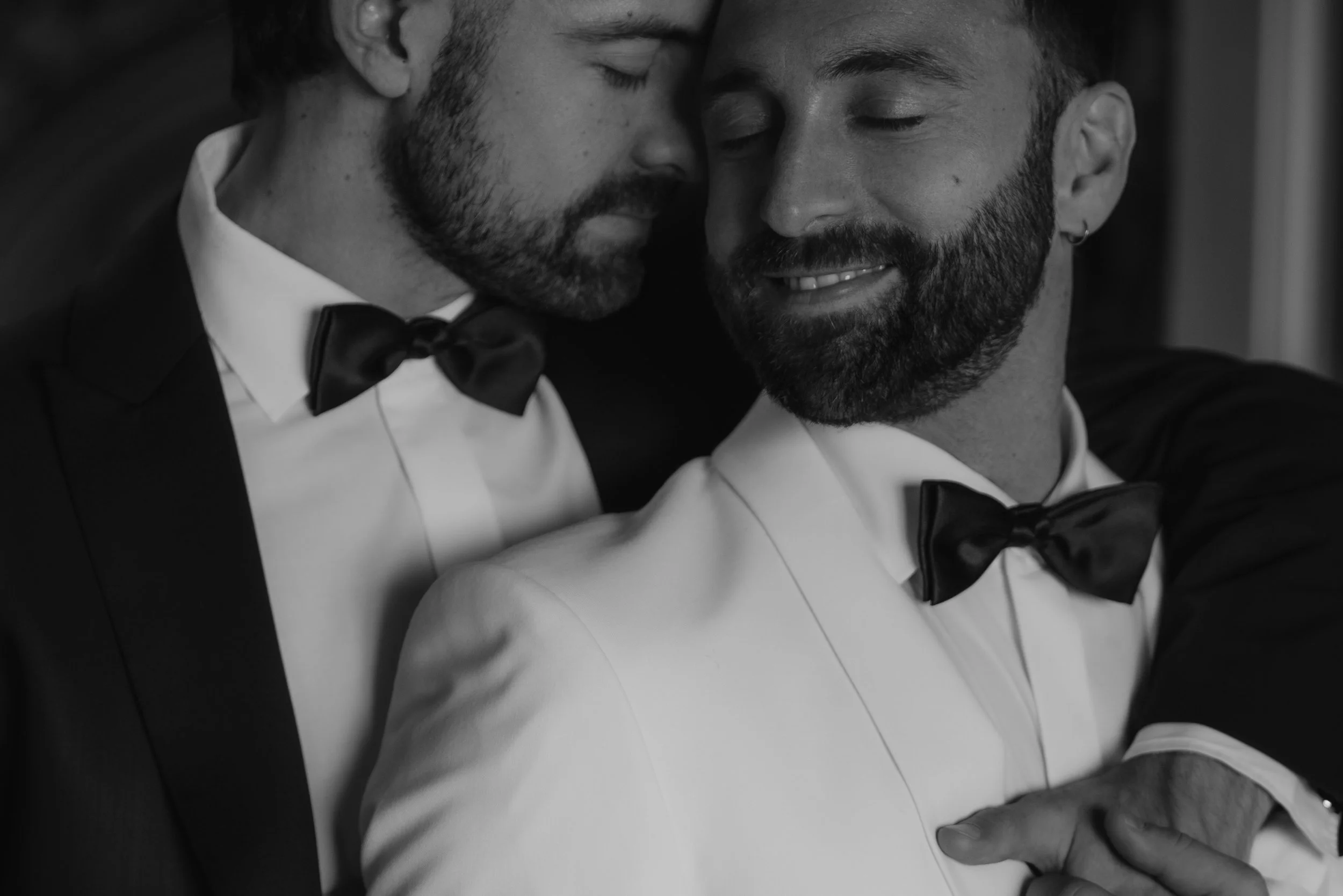 Zwei Männer in formeller Kleidung, die sich umarmen, mit geschlossenen Augen und einem Ausdruck von Glück und Liebe, in Schwarz-Weiß-Fotografie.