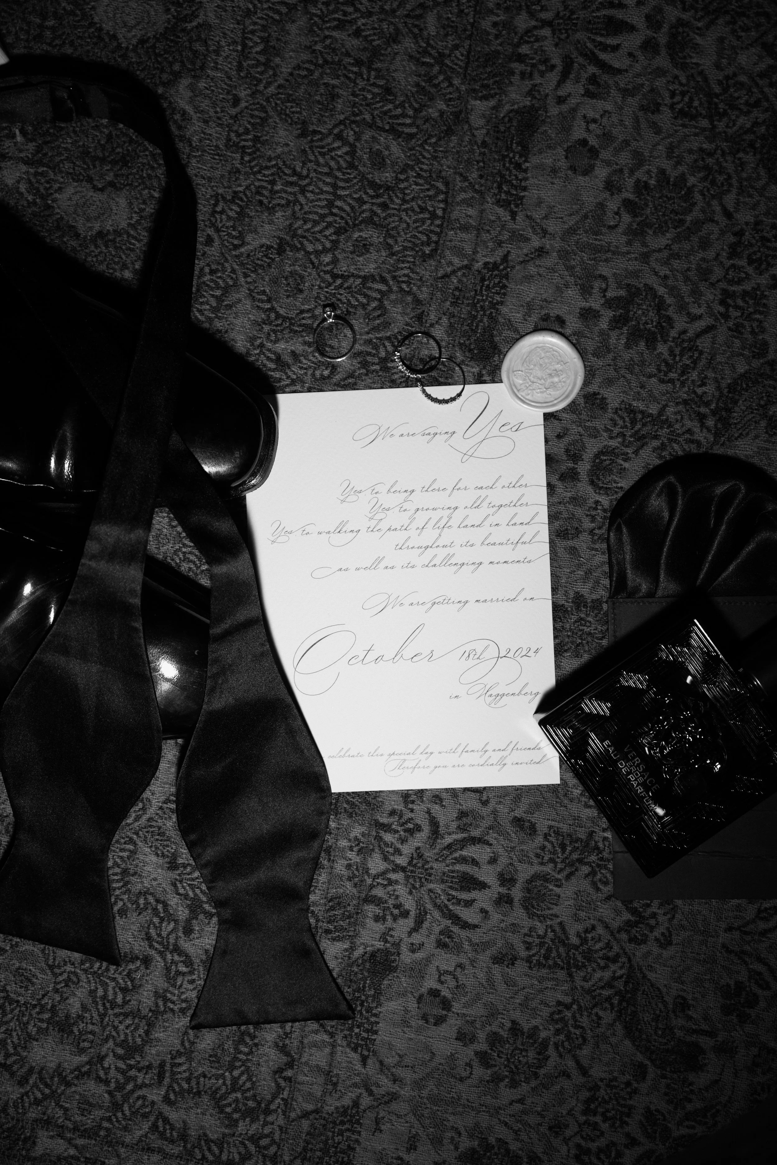 Ein schwarz-weiß Bild zeigt eine Einladung zur Hochzeit, einen schwarzen Schuh mit Schleife, zwei Ringe und eine Spitze Parfüm, alle auf einem Teppichboden.