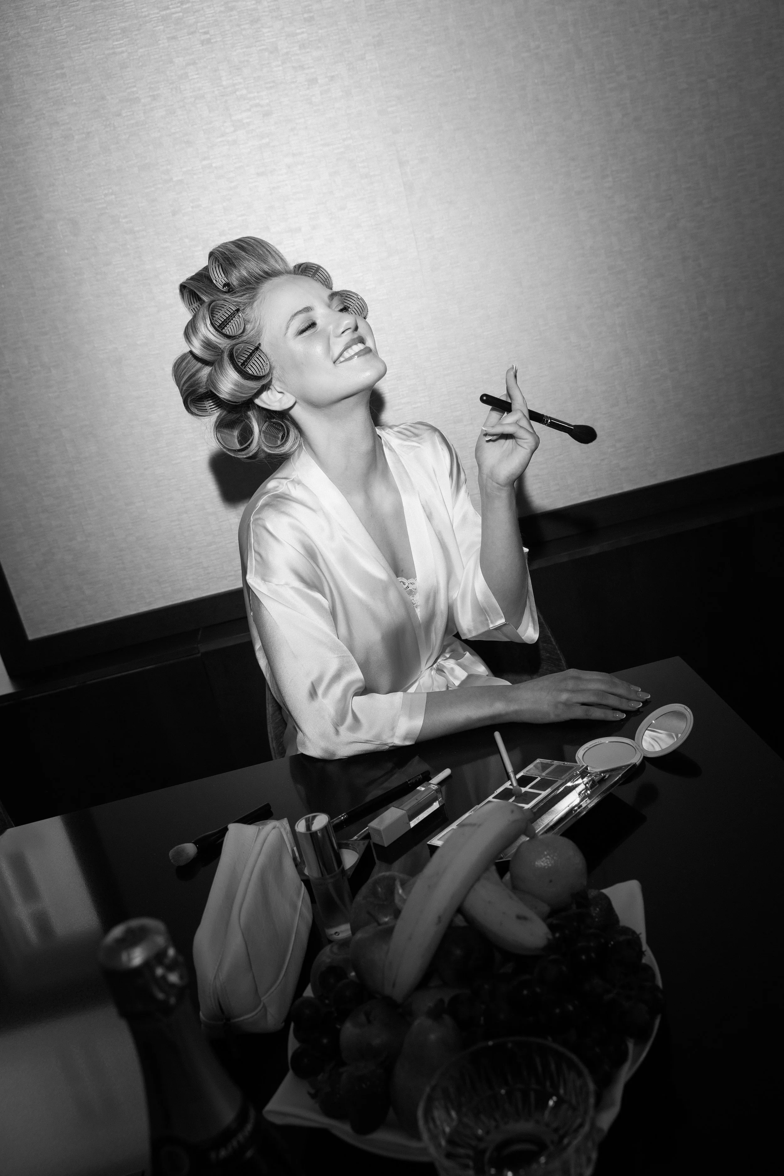 Schwarz-Weiß-Foto einer lachenden jungen Frau mit Wicklern im Haar, die sich im Badezimmer schminkt. Sie trägt eine Satin-Robe und sitzt an einem Tisch mit Make-up-Produkten und Fruchtkorb.