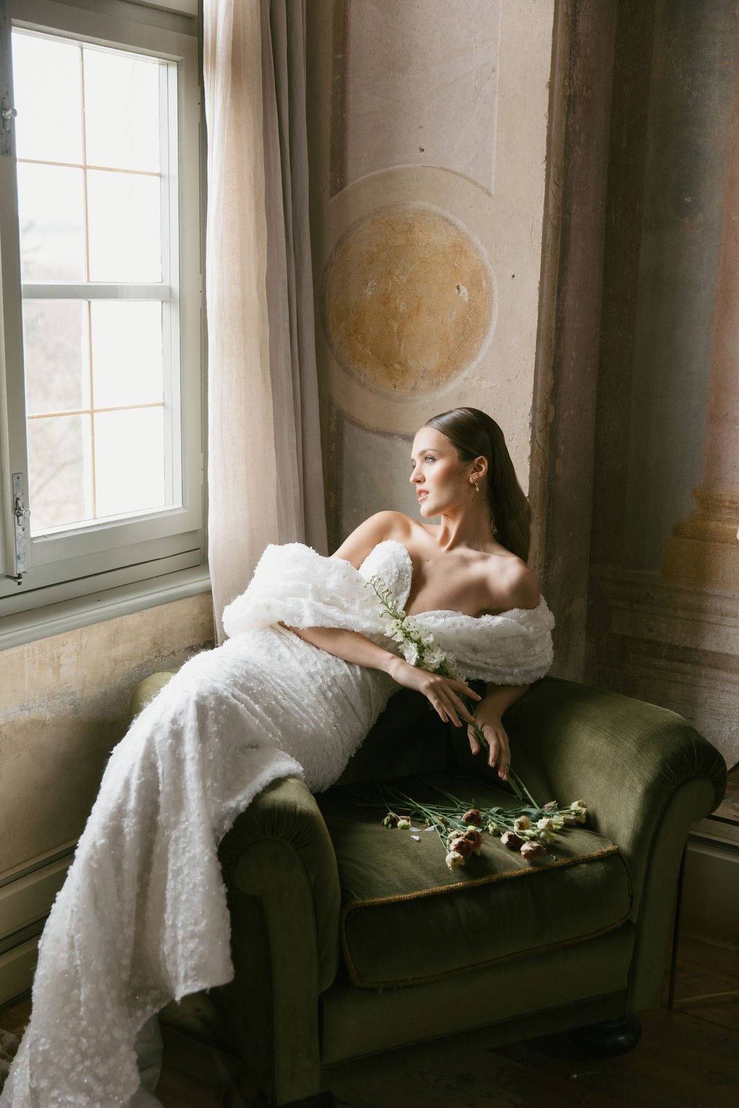 Frau in weißem Brautkleid liegt auf einer grünen Couch vor einem Fenster und blickt nach draußen, mit Blumen in der Hand, in einer historischen, künstlerisch dekorierten Umgebung.