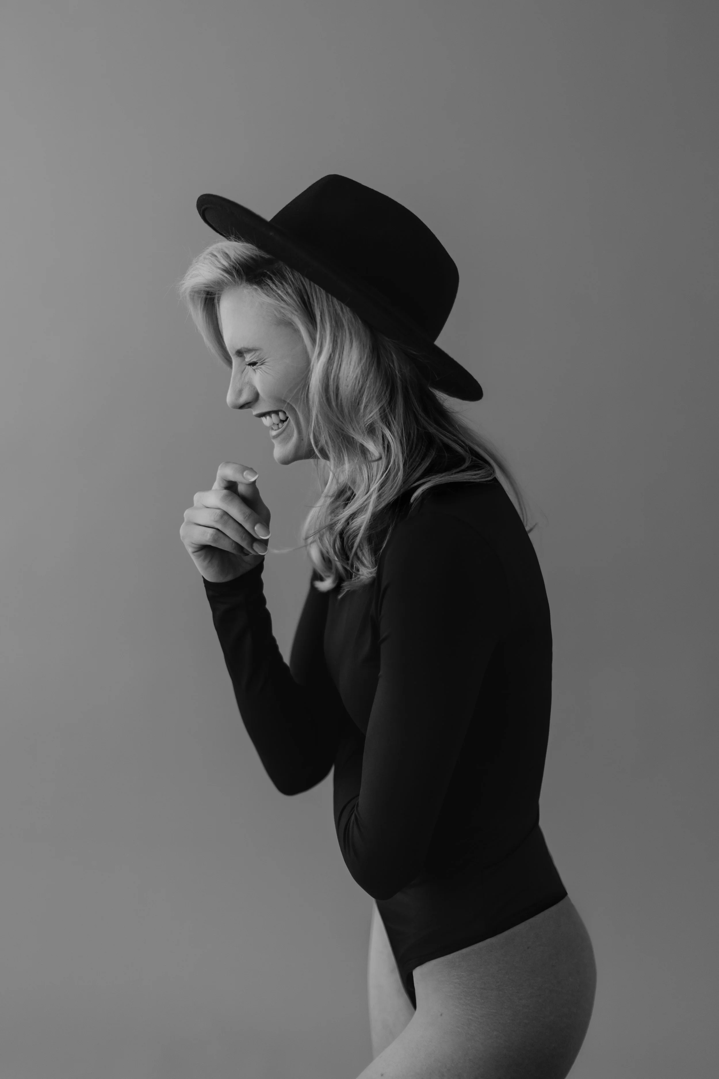 Schwarz-weiß-Fotografie einer lachenden Frau mit Hüftschwung, Sonnenbrille, schwarzem Hut und schwarzem langärmligem Oberteil, die seitlich steht.