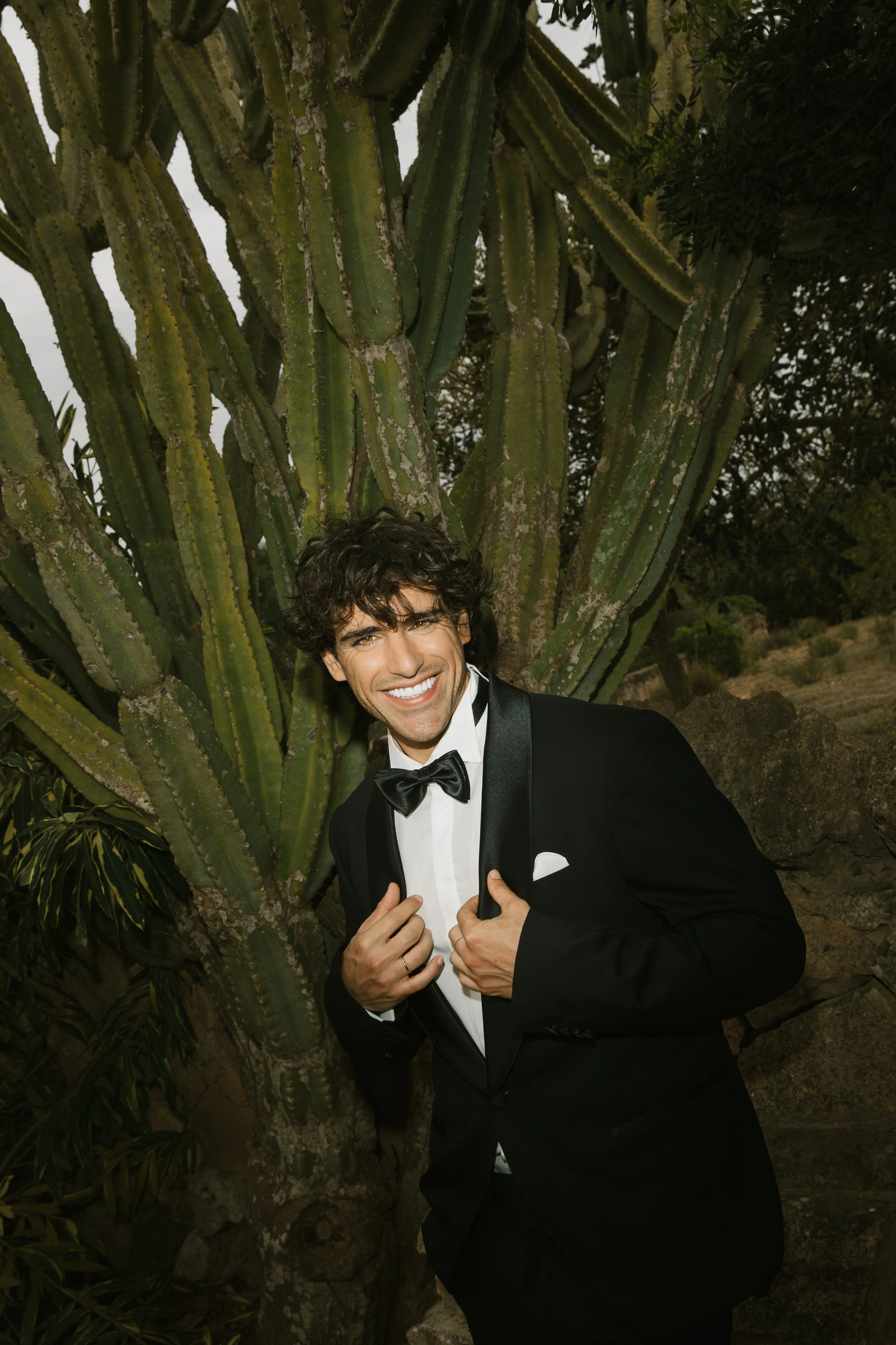 Ein lachender Mann in einem schwarzen Abendanzug mit Fliege posiert vor einer großen Kaktee im Freien.