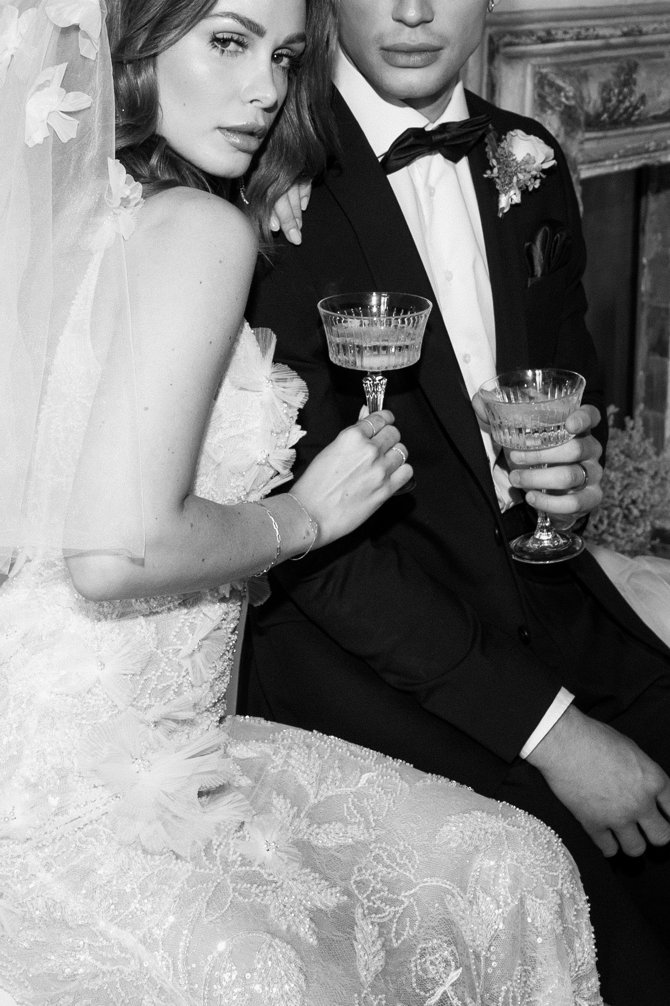 Ein Hochzeitsbild mit einem Brautpaar in formeller Kleidung, die Braut trägt ein aufwendiges Spitzenkleid und eine Schleier, der Bräutigam trägt einen schwarzen Anzug mit Fliege. Beide halten Kristallgläser mit Getränk, sitzen nebeneinander in einem 