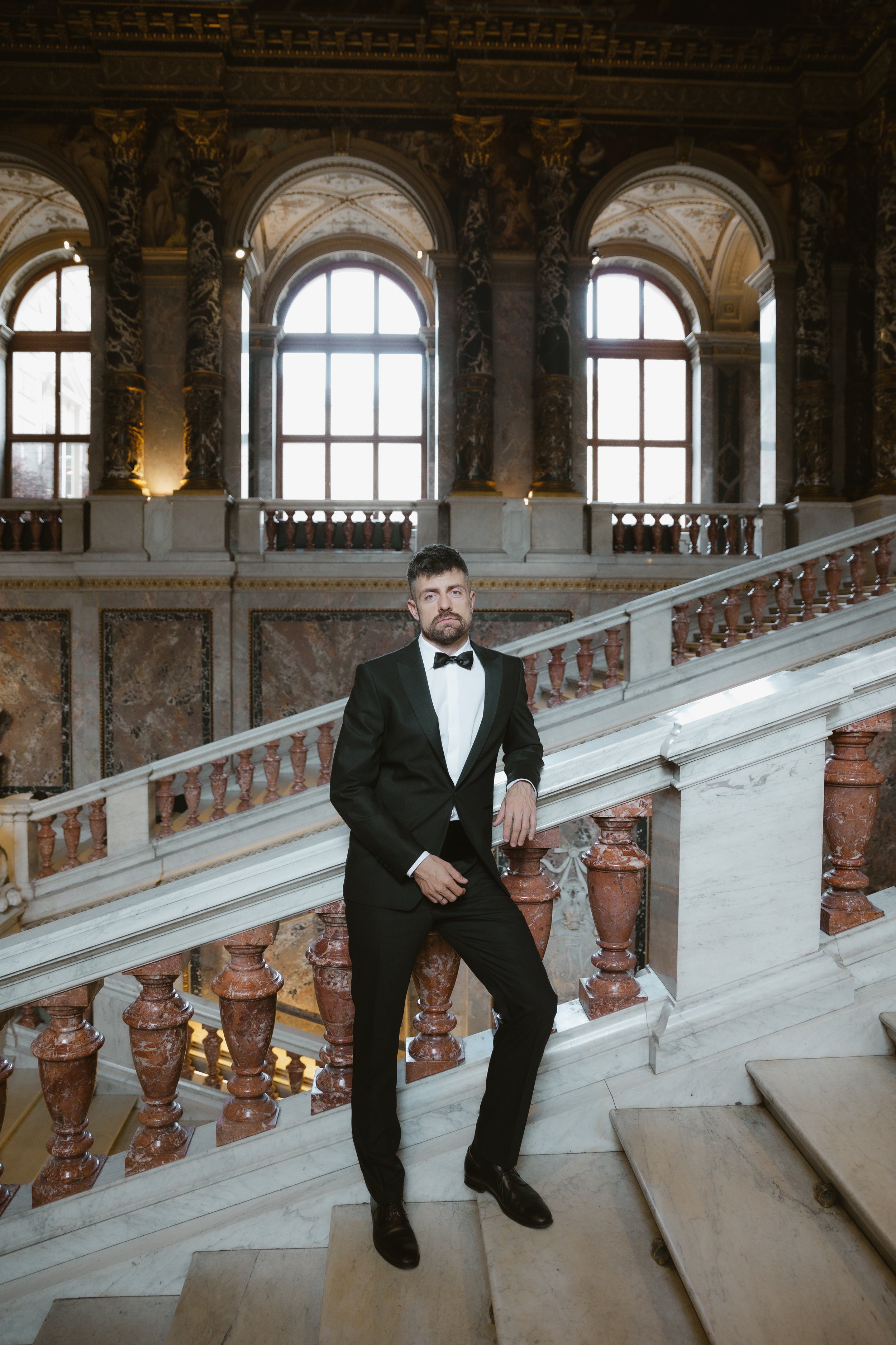 Ein Mann in Smoking steht auf einer Marmortreppe in einem prunkvollen, goldverzierten Saal mit hohen Fenstern und barocken Säulen.