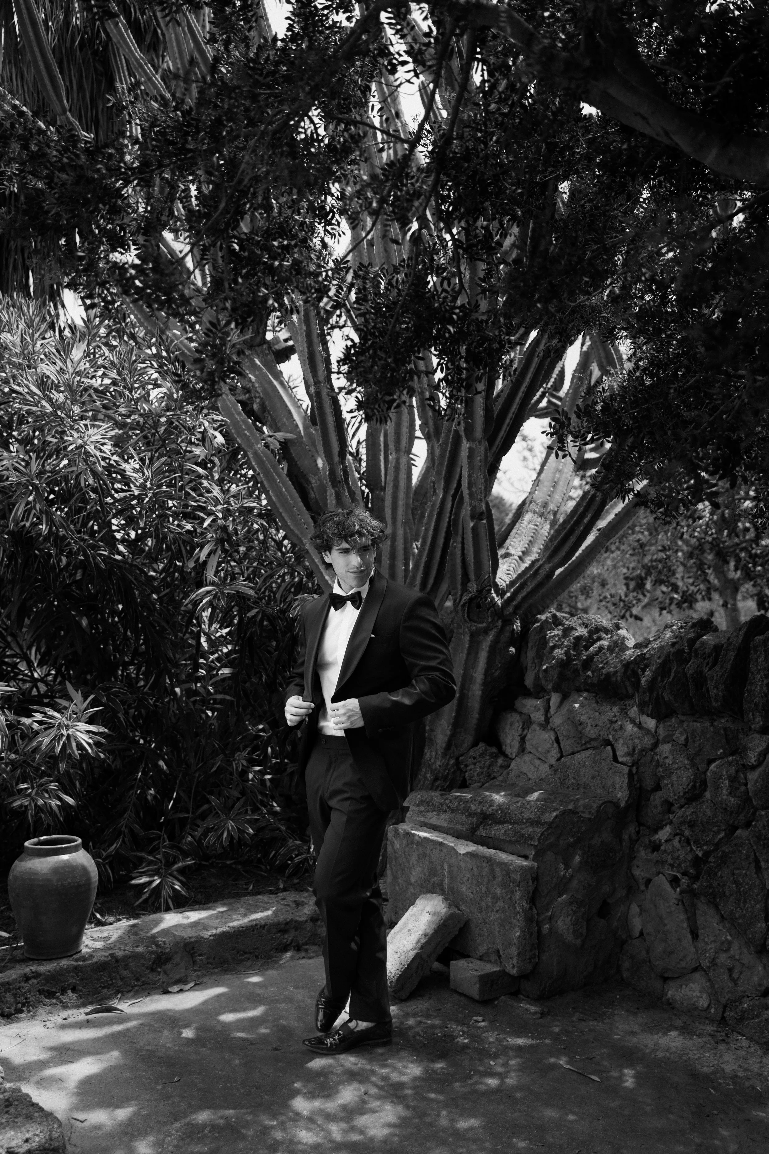 Ein junger Mann in einem schwarzen Smoking mit Fliege posiert vor einem großen Baum und einer Steinmauer, umgeben von Pflanzen, in Schwarz-Weiß.