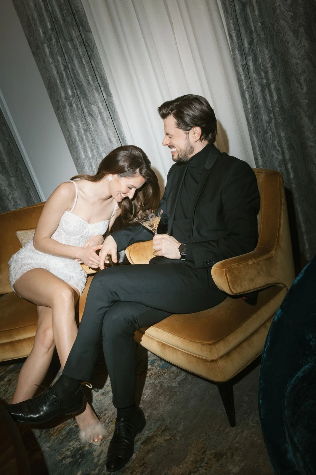 Ein Paar sitzt lachend auf einem goldenen Sofa. Die Frau trägt ein weißes, ärmelloses Kleid und der Mann einen schwarzen Anzug. Sie scheinen Spaß zu haben und halten sich an den Händen. Im Hintergrund sind Vorhänge zu sehen.