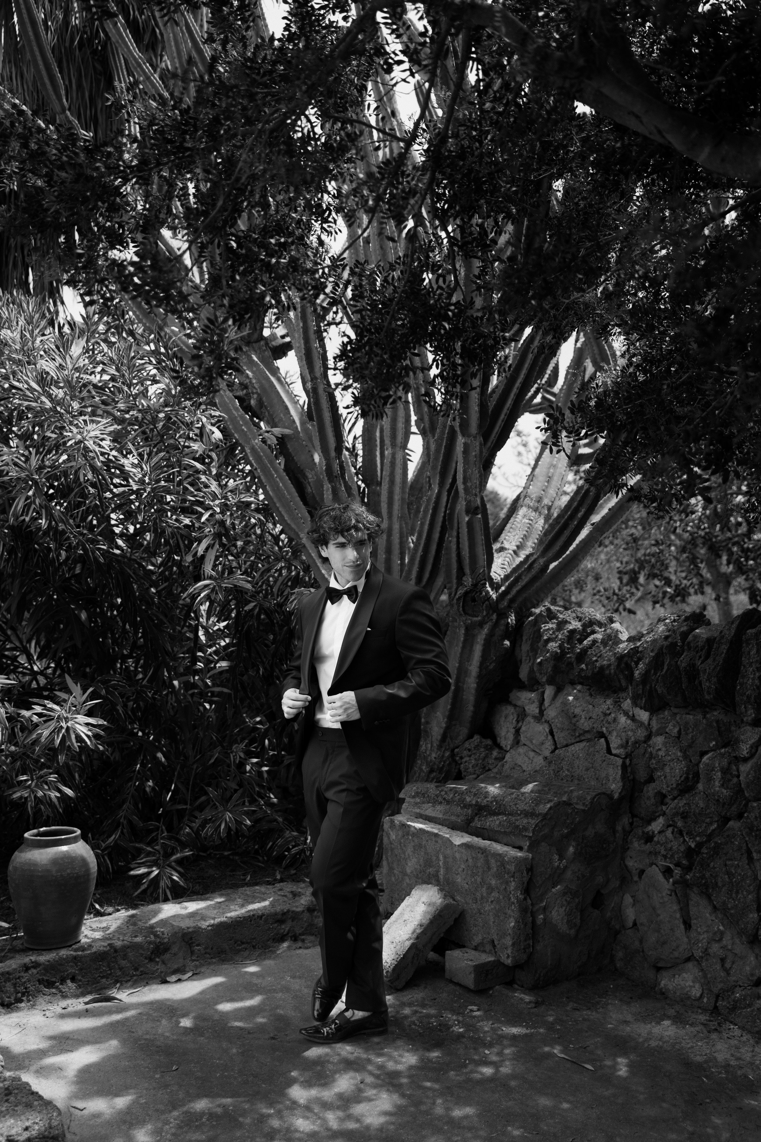 Ein Jugendlicher in einem schwarzen Anzug mit Fliege steht vor einem Baum in einem Garten mit Steinen und einem Keramiktopf.