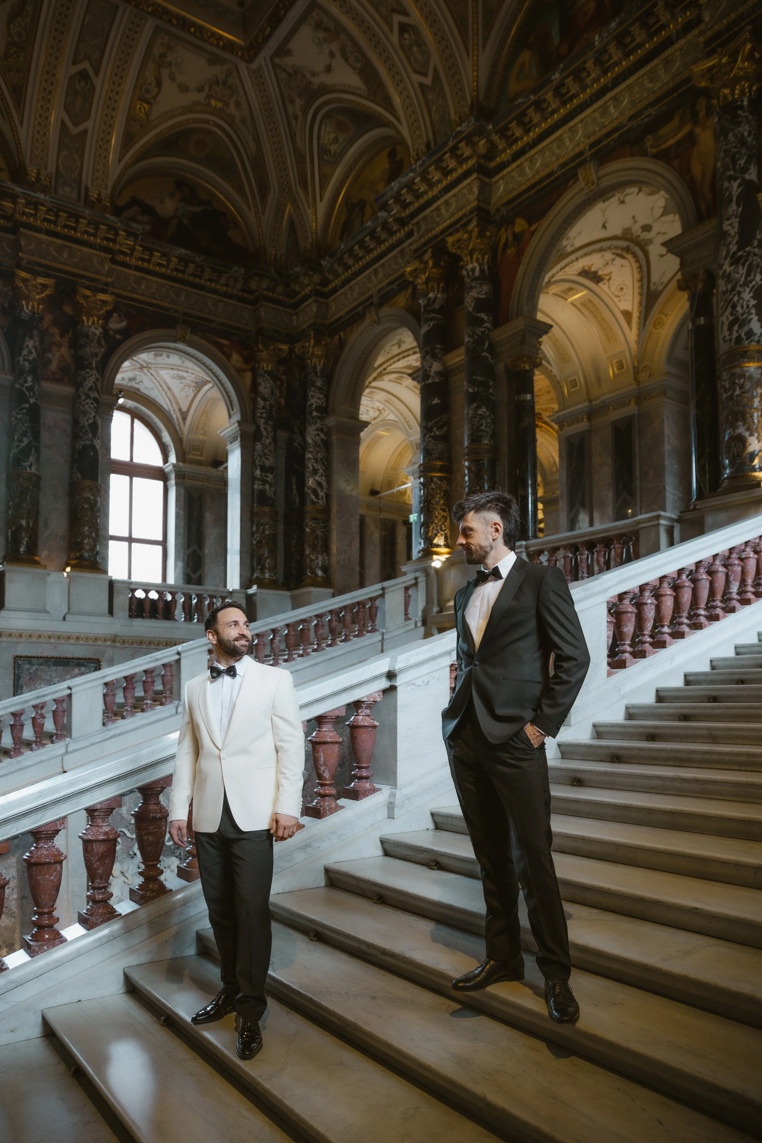 Zwei Männer in schicken Anzügen auf einer Prunktreppe in einem opulenten, barocken Innenraum.