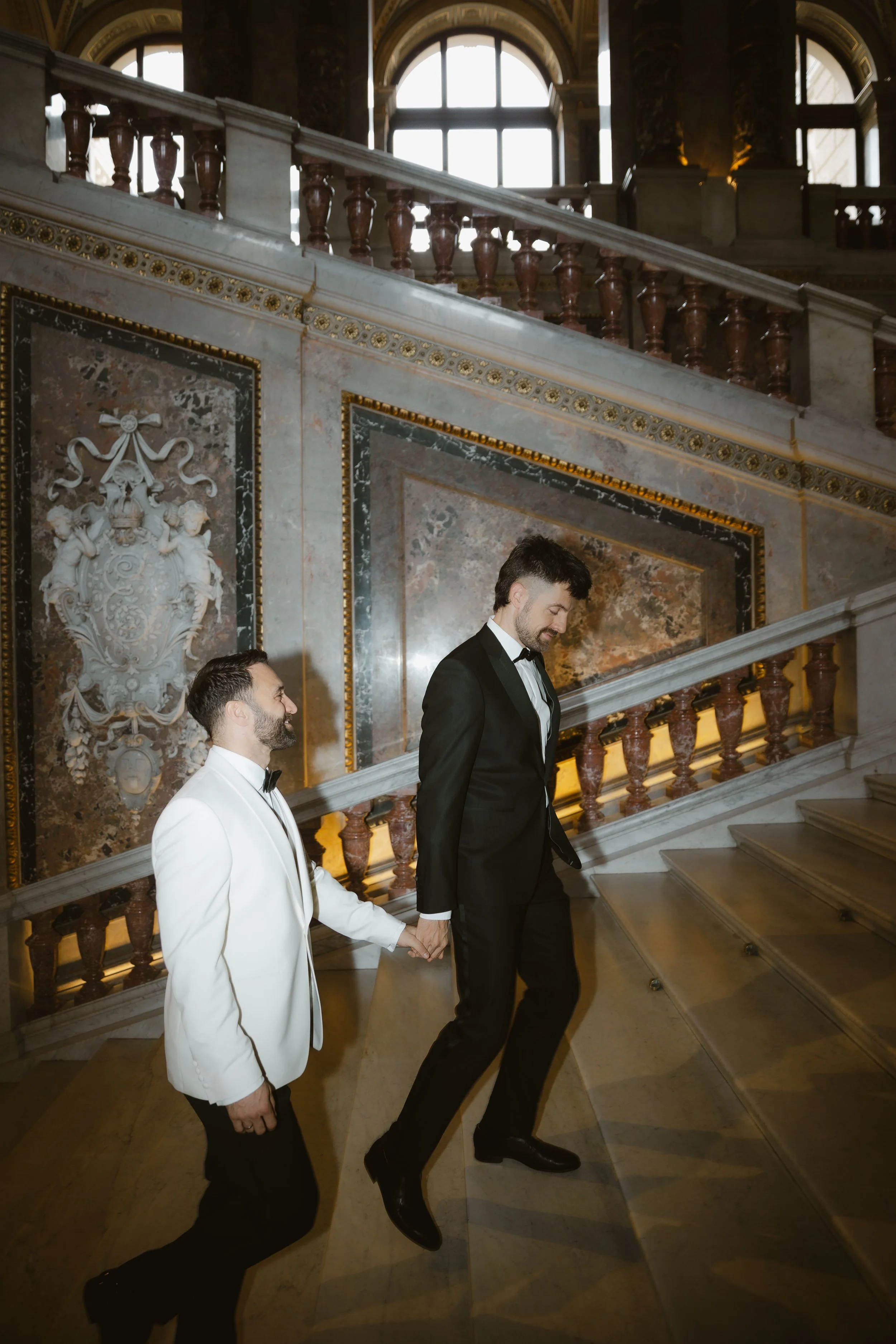 Zwei Männer in formeller Kleidung, einer in weiß und einer in schwarz, steigen die Treppe in einem eleganten, historischen Raum.