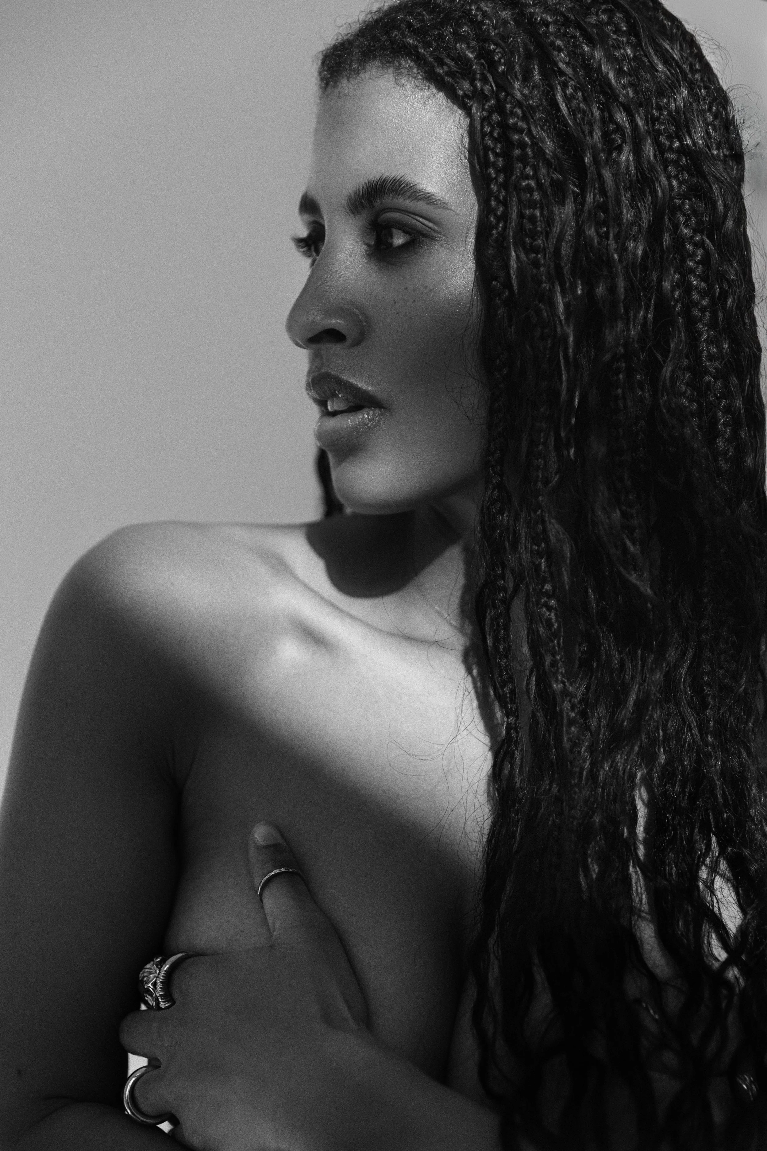 Schwarz-weiß-Fotografie einer Frau, die sich selbst mit einer Hand am Oberkörper berührt, mit lange, strukturierte Haare und nachdenklichem Blick nach links.