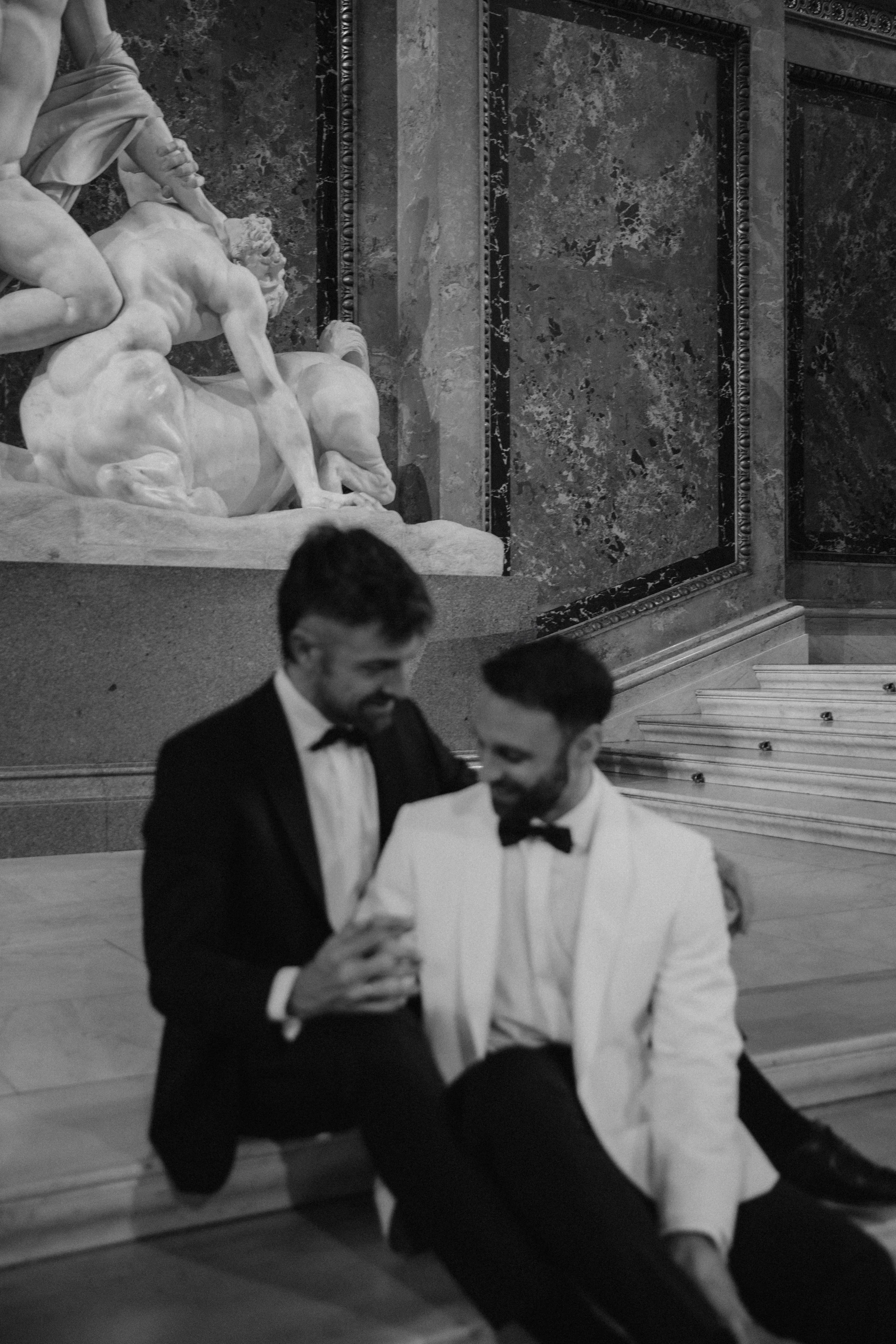 Zwei Männer in formeller Kleidung, die in einem Museum sitzen und auf ein Smartphone schauen, im Hintergrund eine Skulptur.
