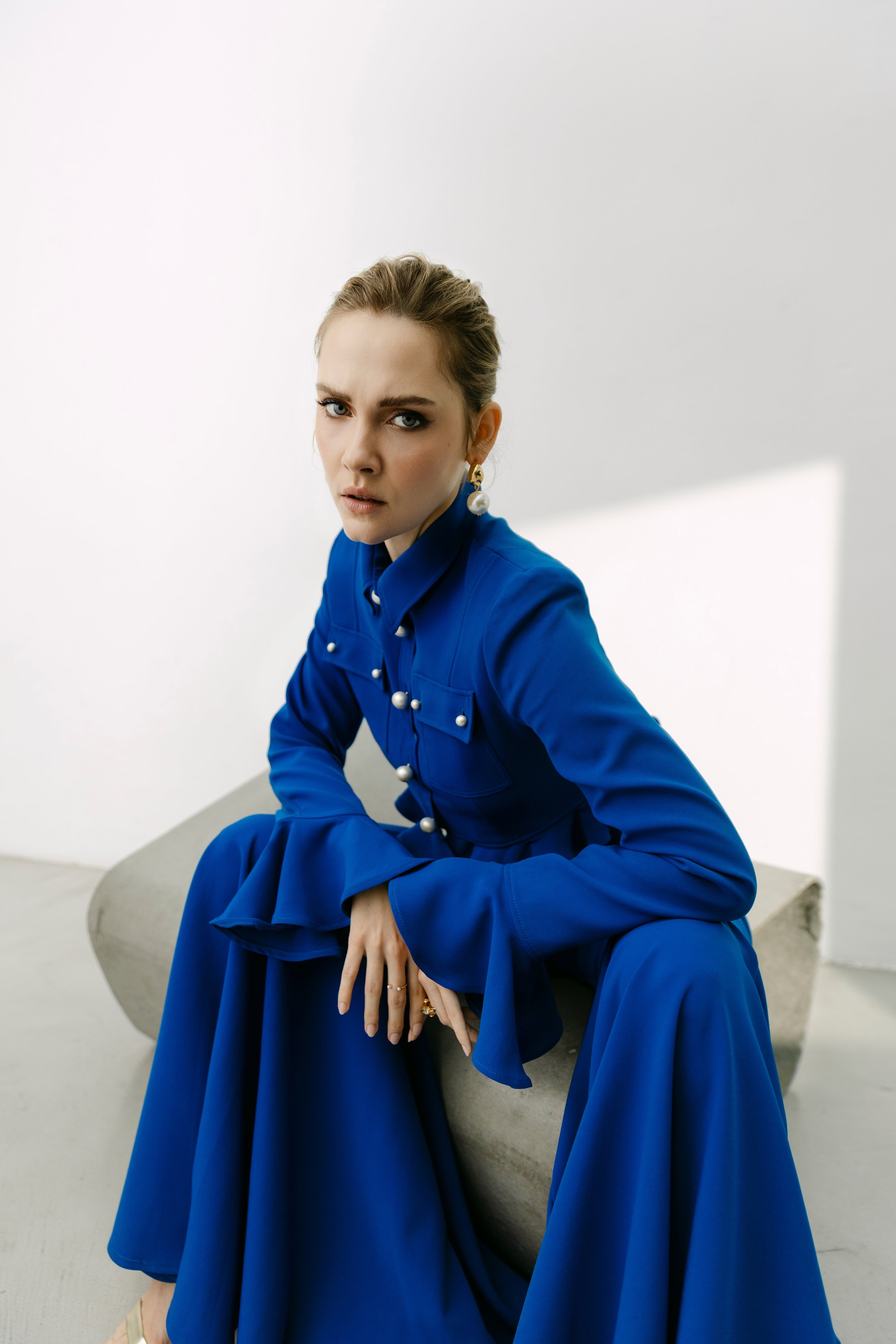 Eine junge Frau in einem eleganten blauen Kleid sitzt auf einer Betonbank vor einem weißen Wand. Sie schaut direkt in die Kamera mit einem ernsten Ausdruck, trägt große Perlenohrringe und eine Perlenkette.
