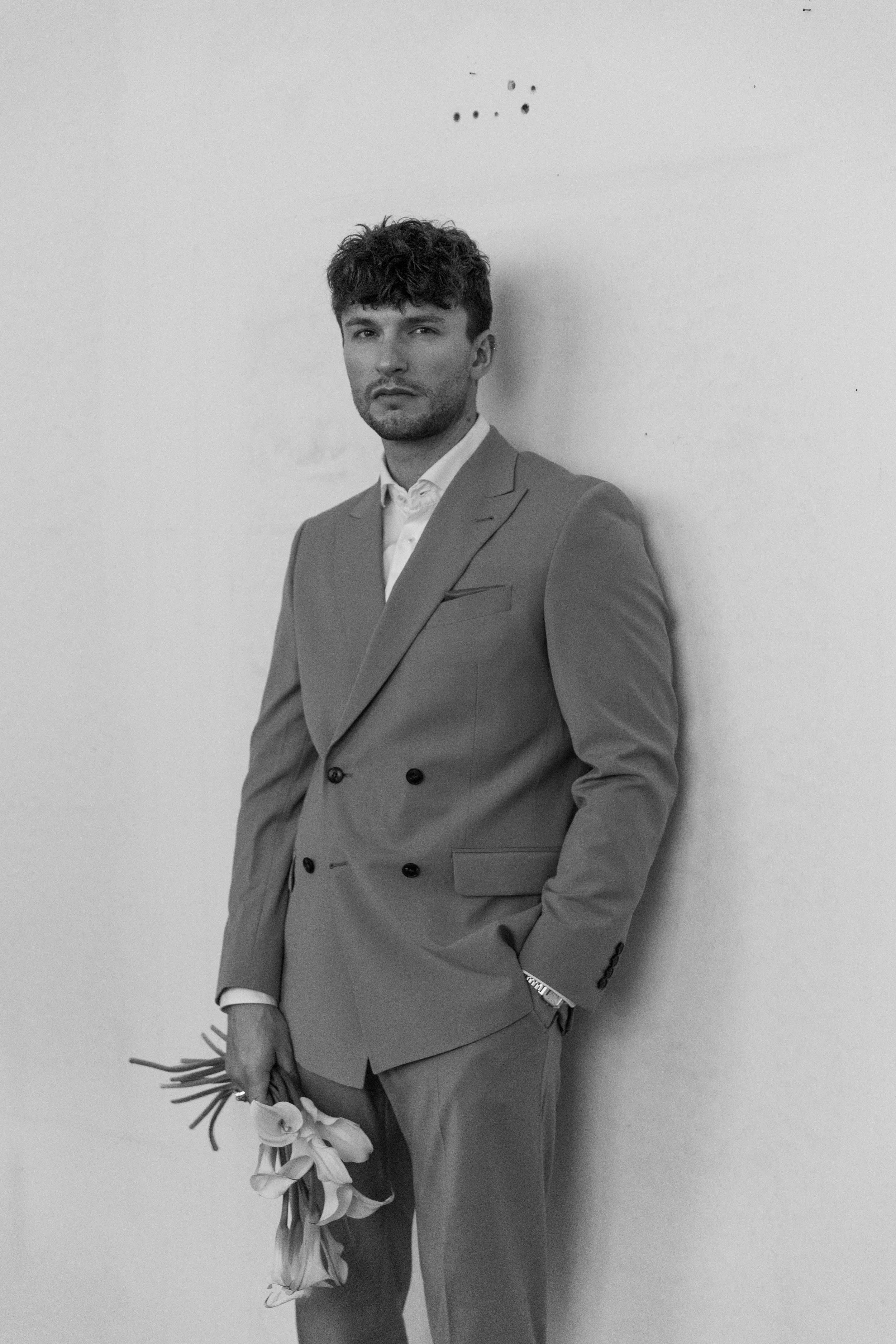 Schwarz-Weiß-Foto eines Mannes in Anzug, der an einer weißen Wand lehnt und eine Tulpe hält.