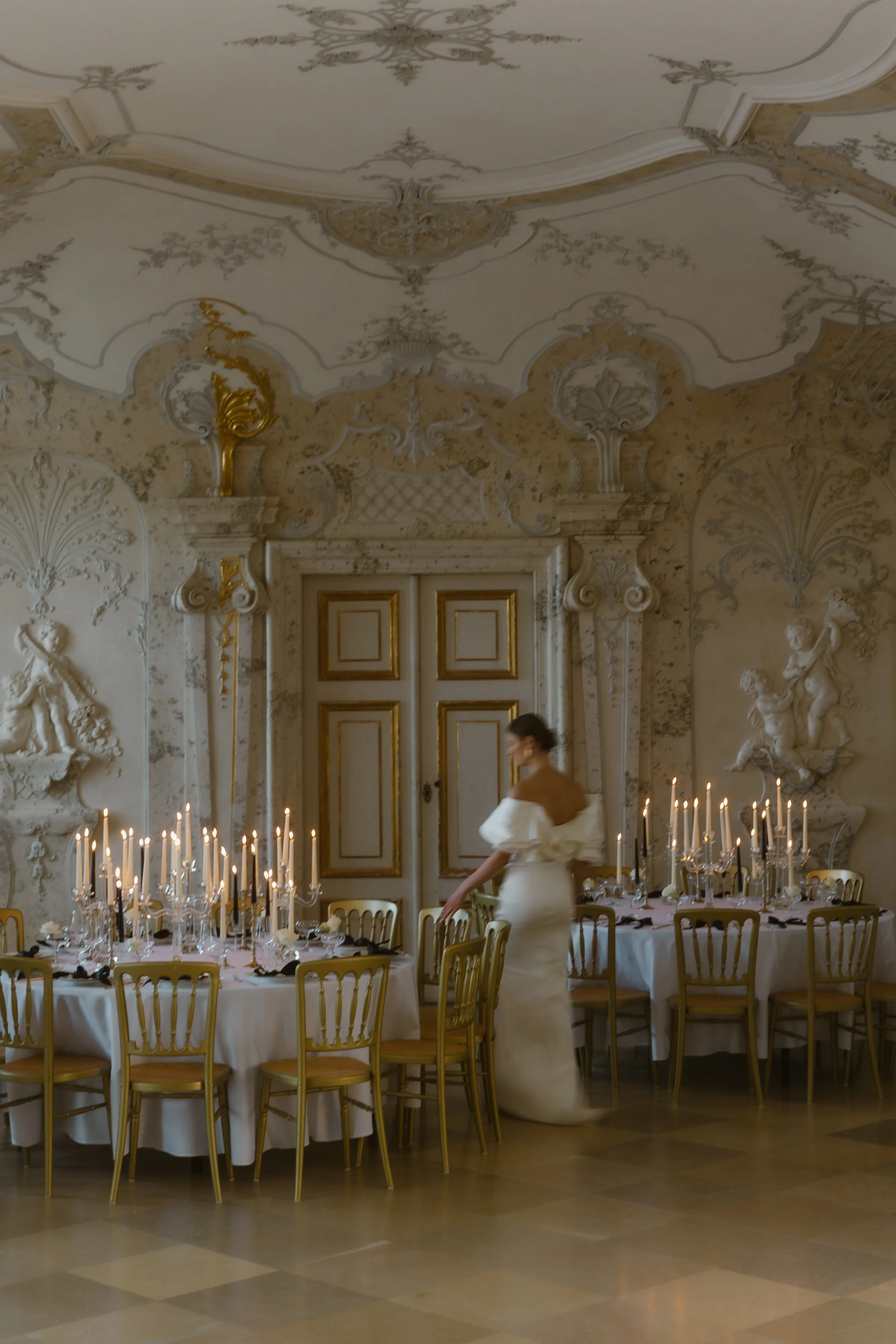 Eine elegante Frau in einem weißen Abendkleid steht in einem prunkvollen, barockartig dekorierten Saal vor einem festlich gedeckten Tisch mit Kerzen.