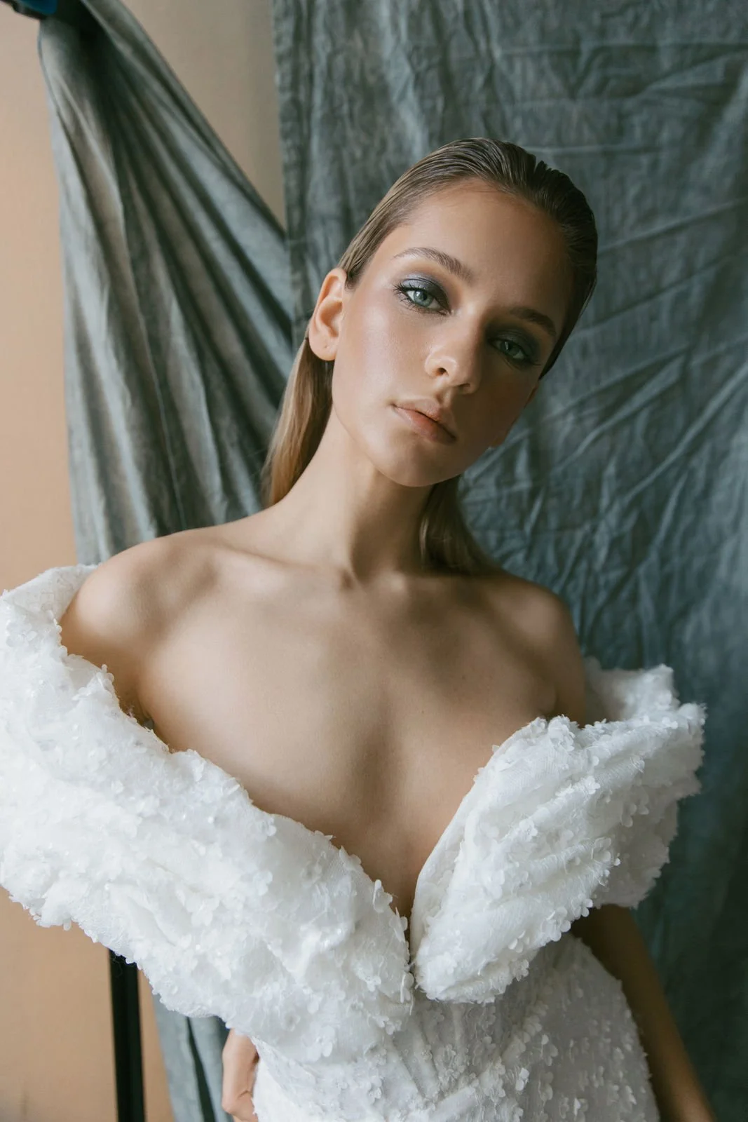 Junge Frau in weißem Kleid mit gepufften Schultern und glatter, langer, blond gefärbter Haare, stilvolles Make-up, blickt ernst in die Kamera, im Hintergrund grauer Vorhang