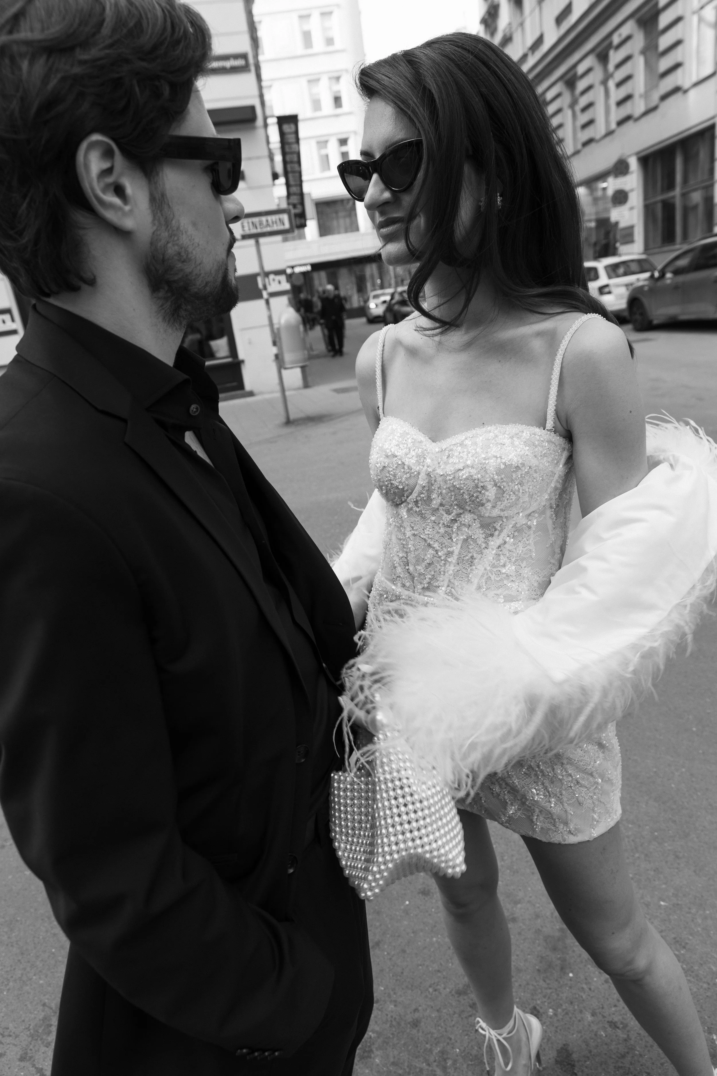 Zwei Personen auf der Straße stehen sich gegenüber, beide tragen Sonnenbrillen. Eine Person trägt ein elegantes Kleid mit Pailletten und hält eine verzierte Handtasche.