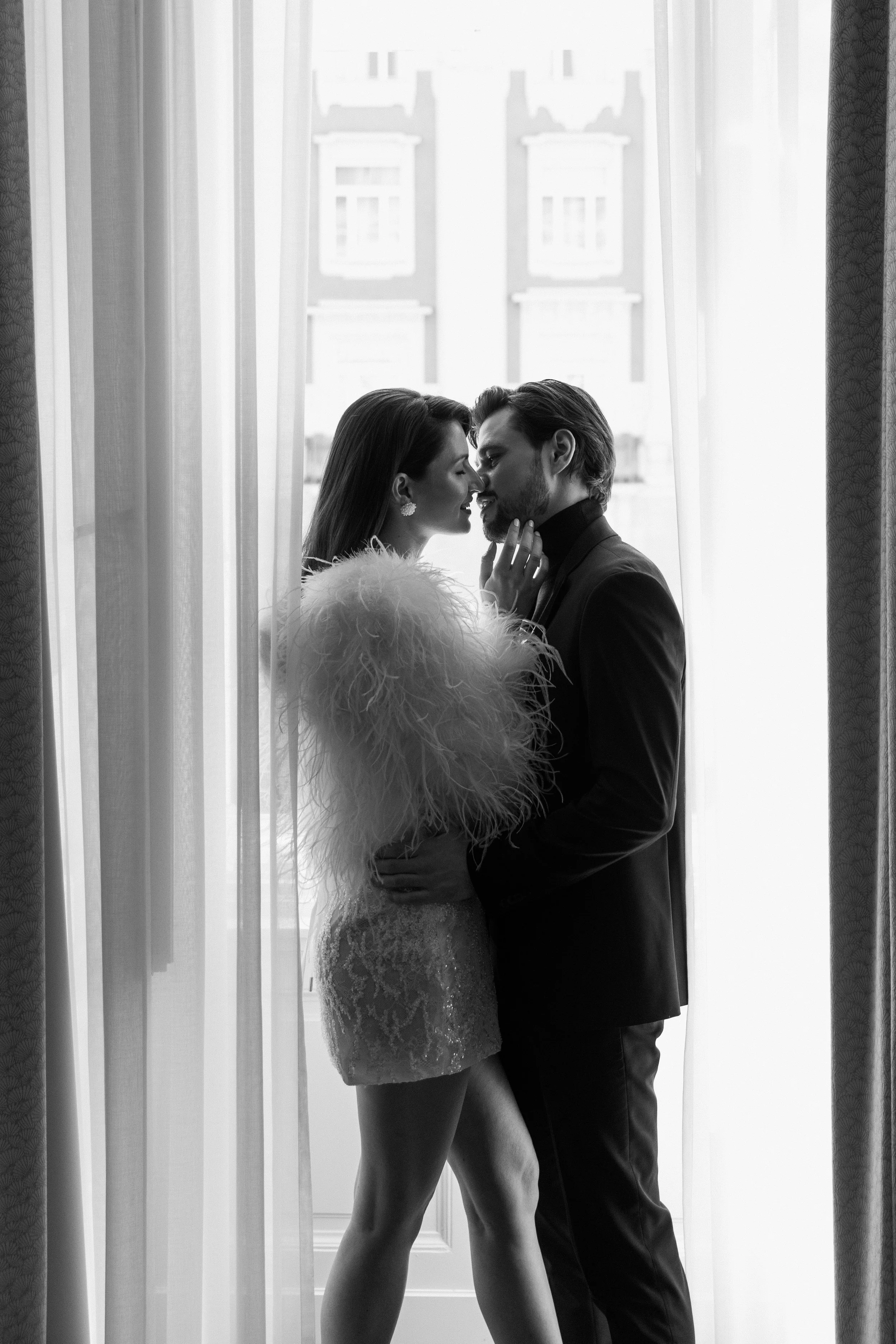 Ein Paar in eleganter Kleidung steht sich vor einem Fenster, ihre Gesichter sehr nah, fast küssend, in einer intimen und romantischen Szene. Es ist eine Schwarz-Weiß-Fotografie.