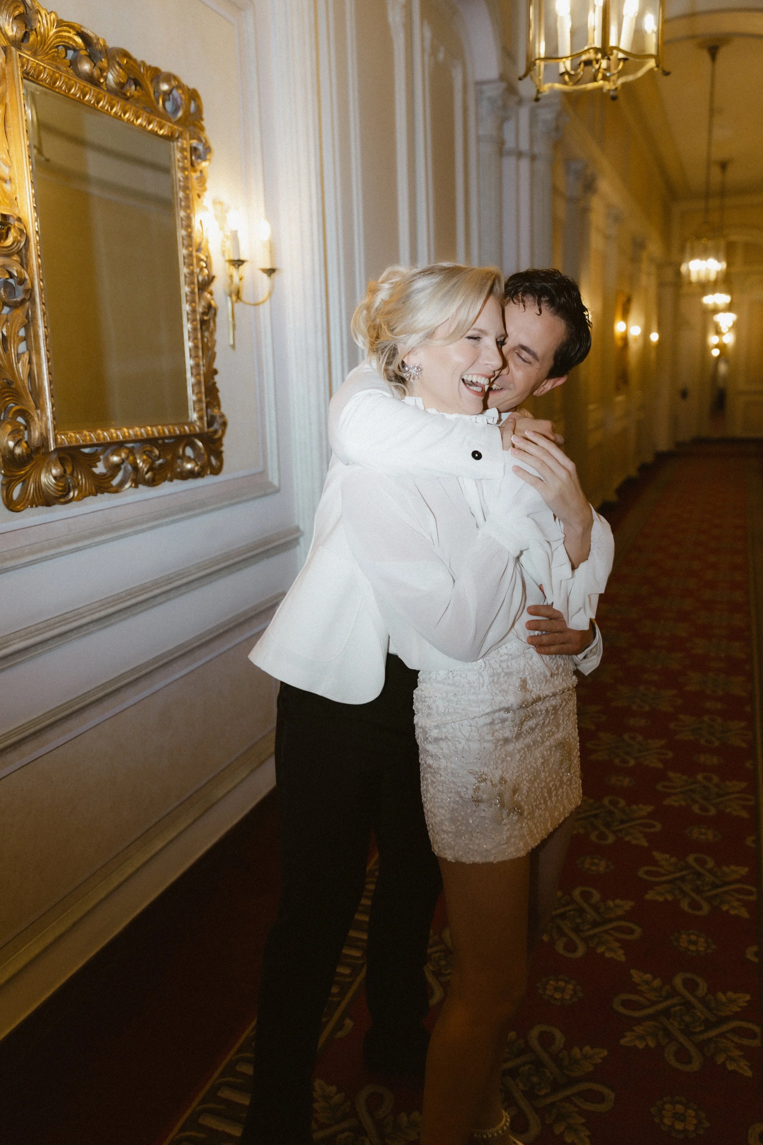Ein lachendes Paar in eleganter Kleidung, umarmend in einem goldverzierten Flur mit Kronleuchtern.