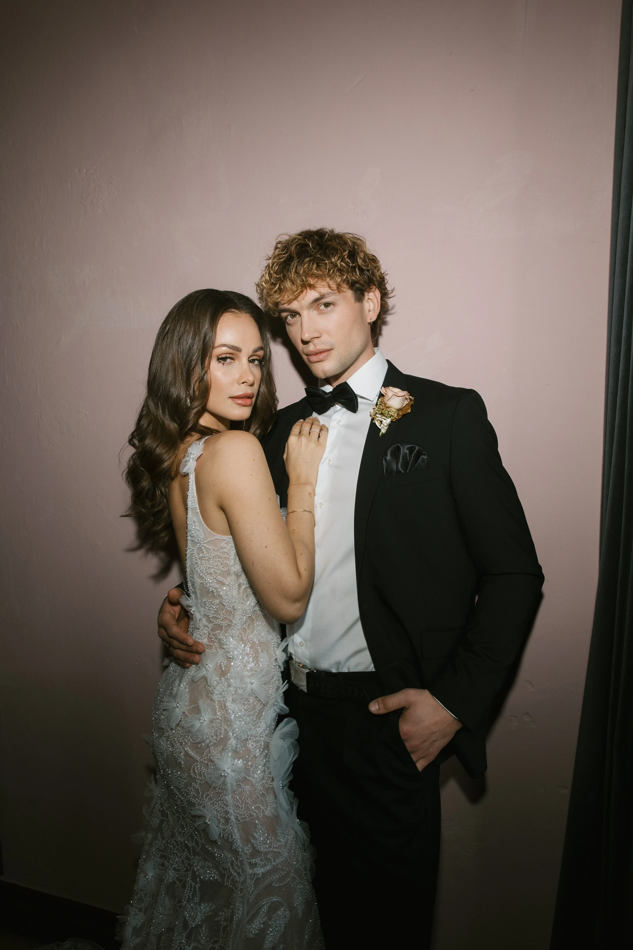 Ein junges Paar in Hochzeitskleidung steht vor einer hellen Wand, das Mädchen trägt ein detailliertes, Spitzehochzeitskleid, der Mann trägt einen schwarzen Anzug mit Fliege, mit einer kleinen Blume am Revers.