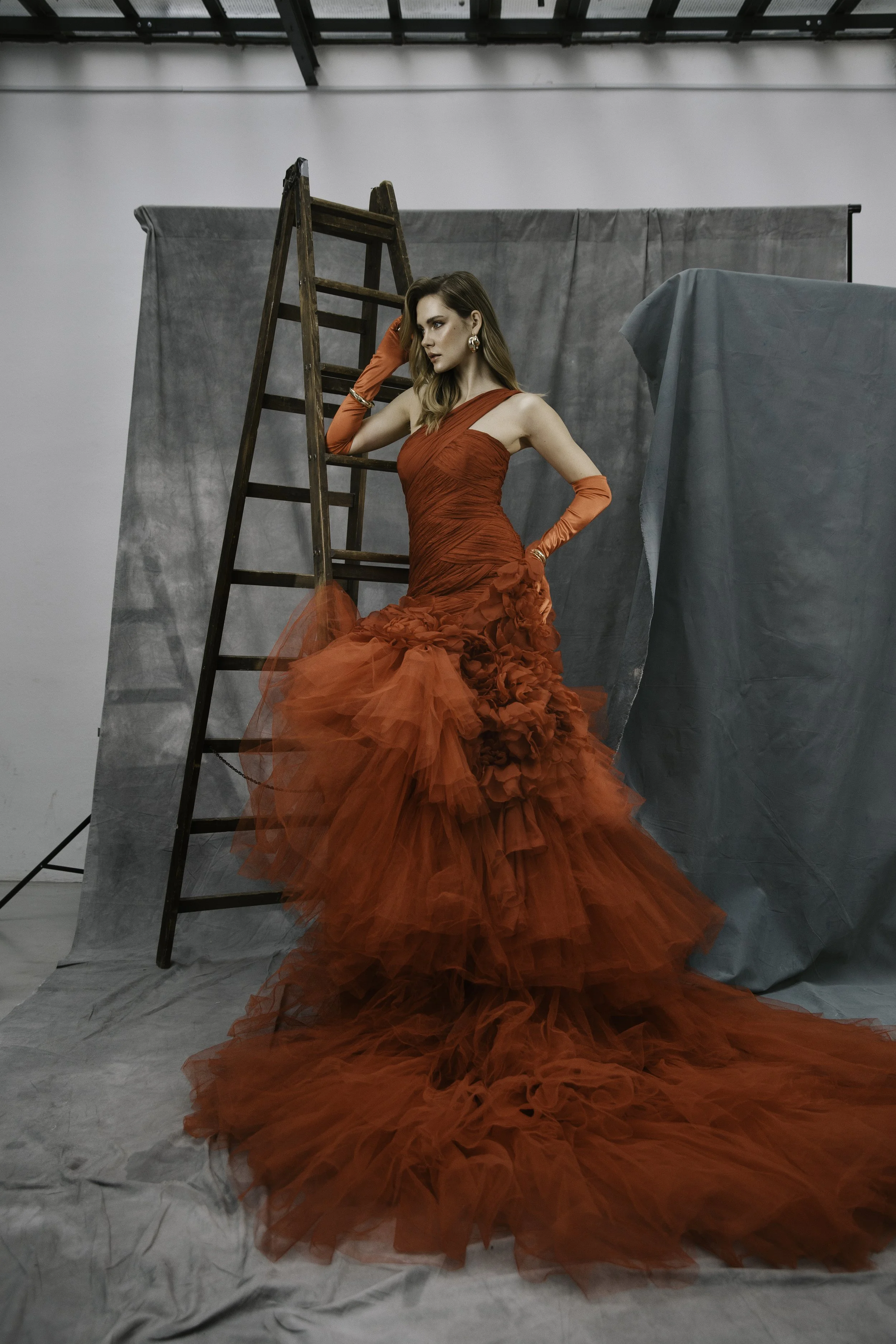 Frau in rotem Abendkleid poseiert vor einer grauen Stoffhintergrund im Studio, neben einer Leiter, entstehendes Mode- oder Fotoshooting.