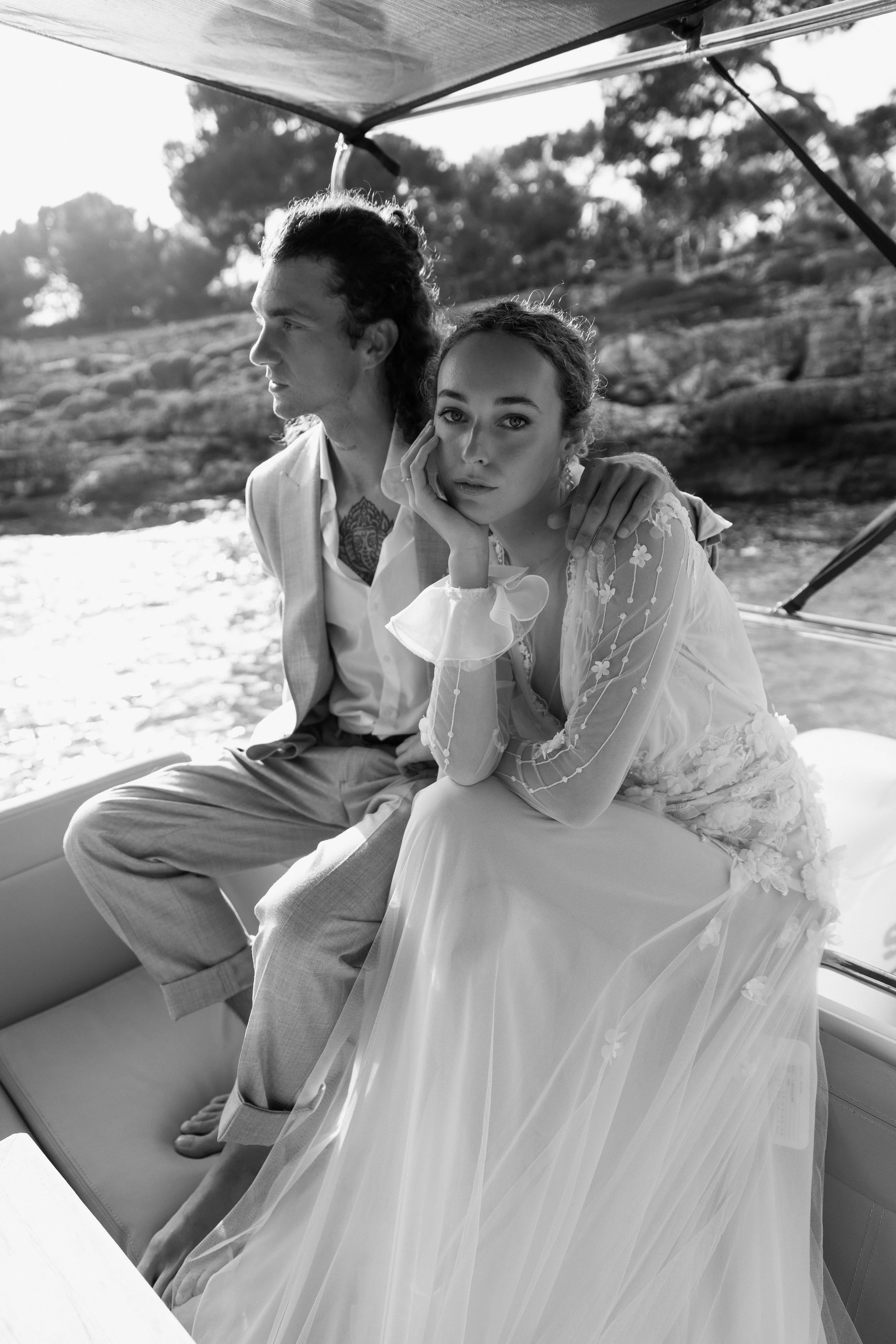 Schwarz-Weiß-Foto eines Paares auf einem Boot, die Frau trägt ein Brautkleid und der Mann einen Anzug. Im Hintergrund sind Bäume und Wasser.