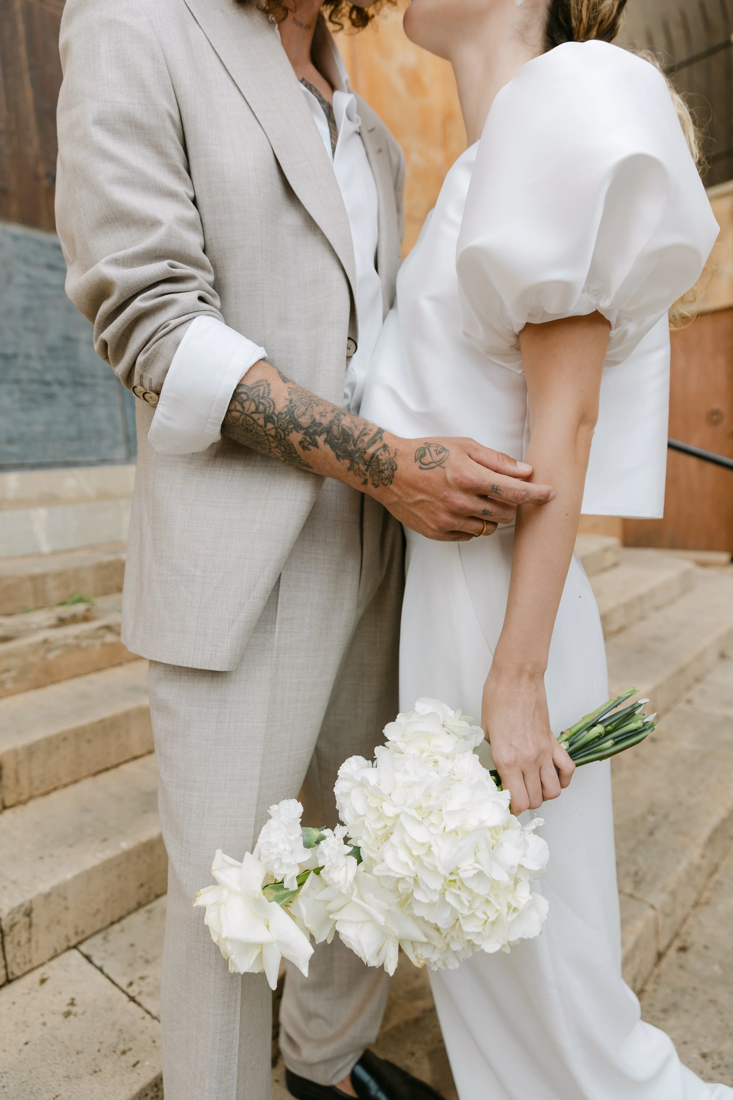 Nahaufnahme des Brautpaares, beide halten einen weißen Blumenstrauß, die Braut trägt ein weißes Kleid mit Puffärmeln, der Bräutigam trägt einen beigen Anzug, sie stehen auf Treppen vor einem Holzhaus.
