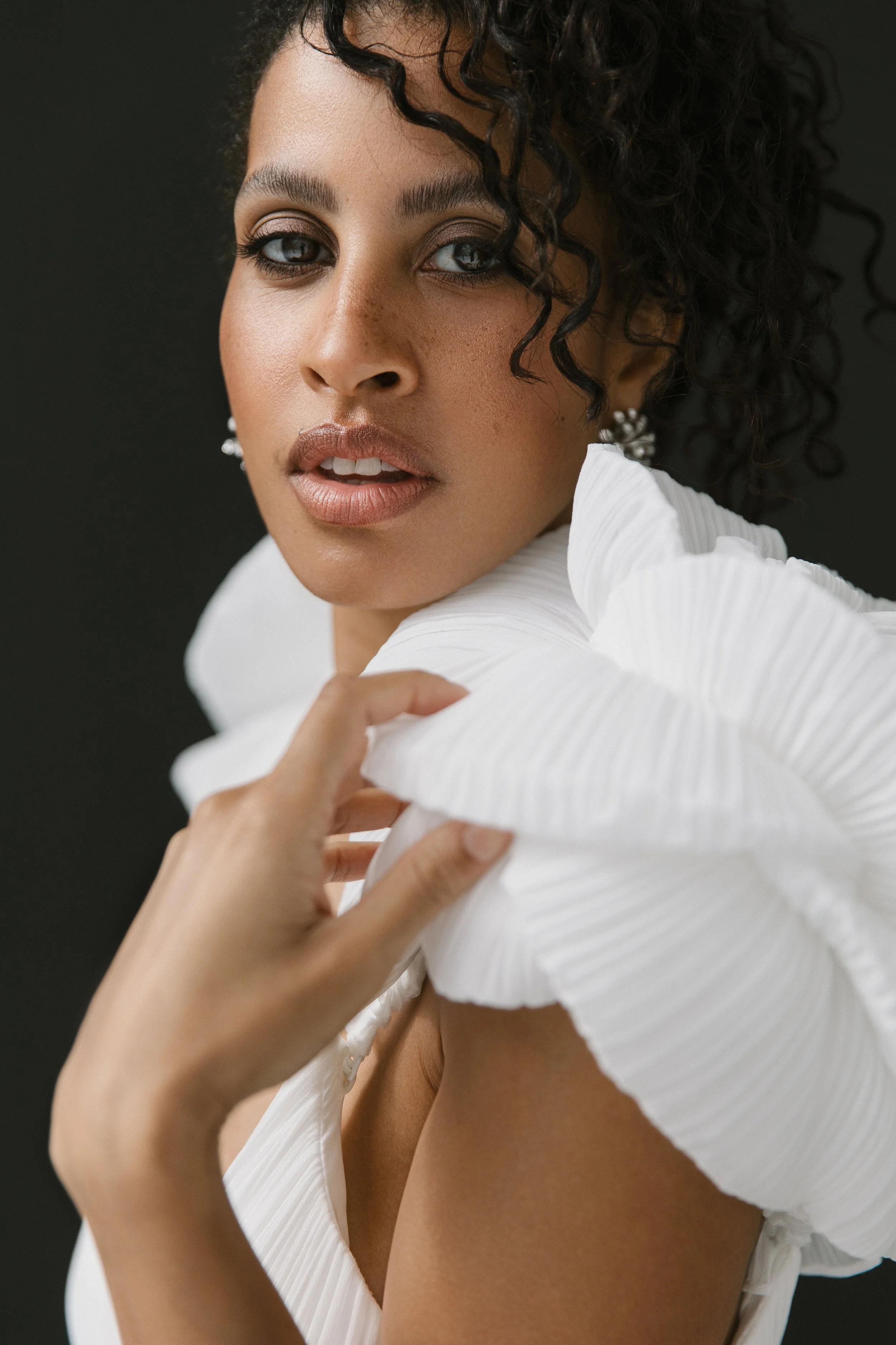 Nahaufnahme einer jungen Frau mit dunklen, lockigen Haaren und hellbraunen Haut, die einen eleganten weißen, gerüschten Kleidungsstück trägt, vor dunklem Hintergrund.