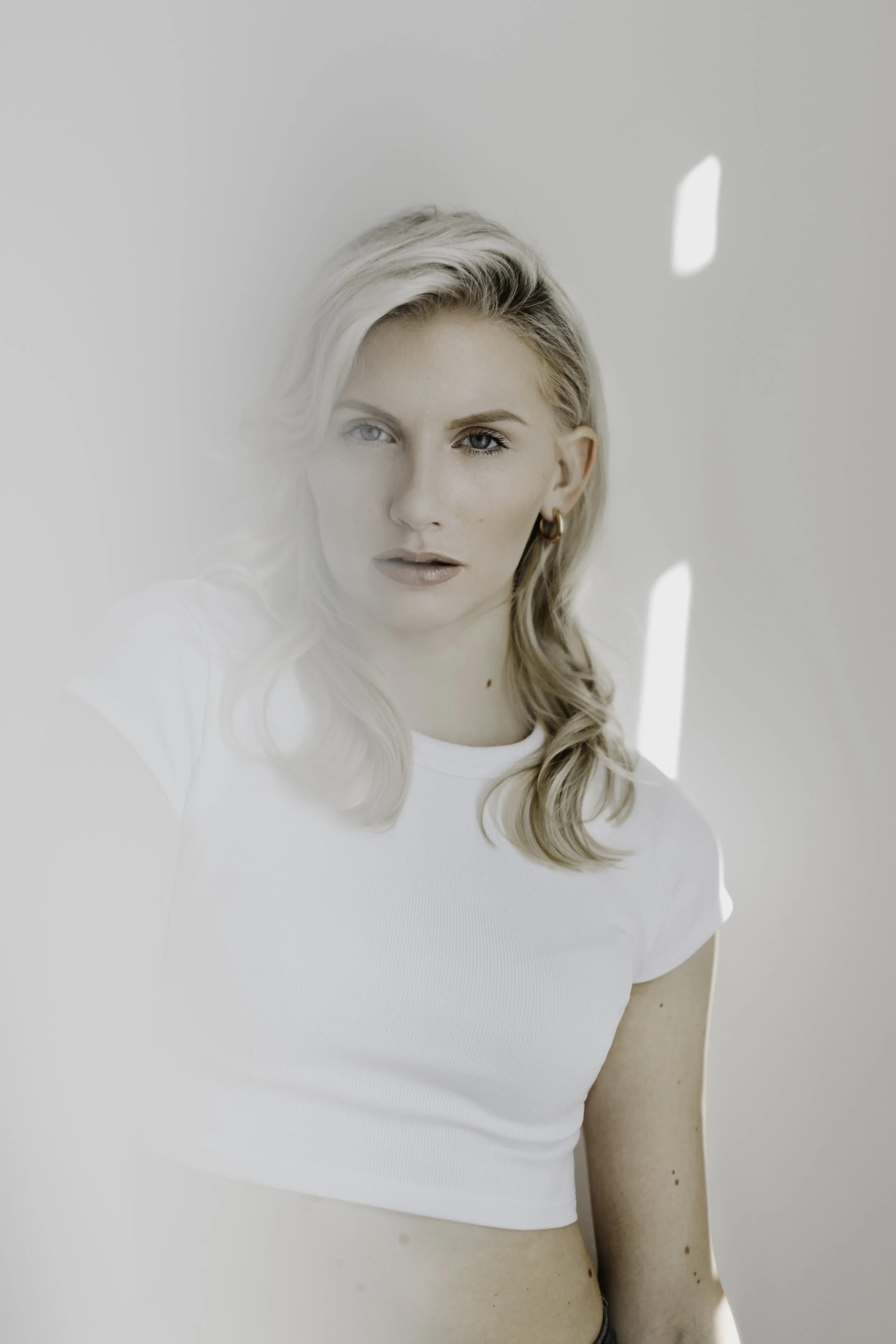 Junge Frau mit blonden, gewellten Haaren, trägt eine weiße T-Shirt und blickt in die Kamera, mit Lichtreflexen im Hintergrund.