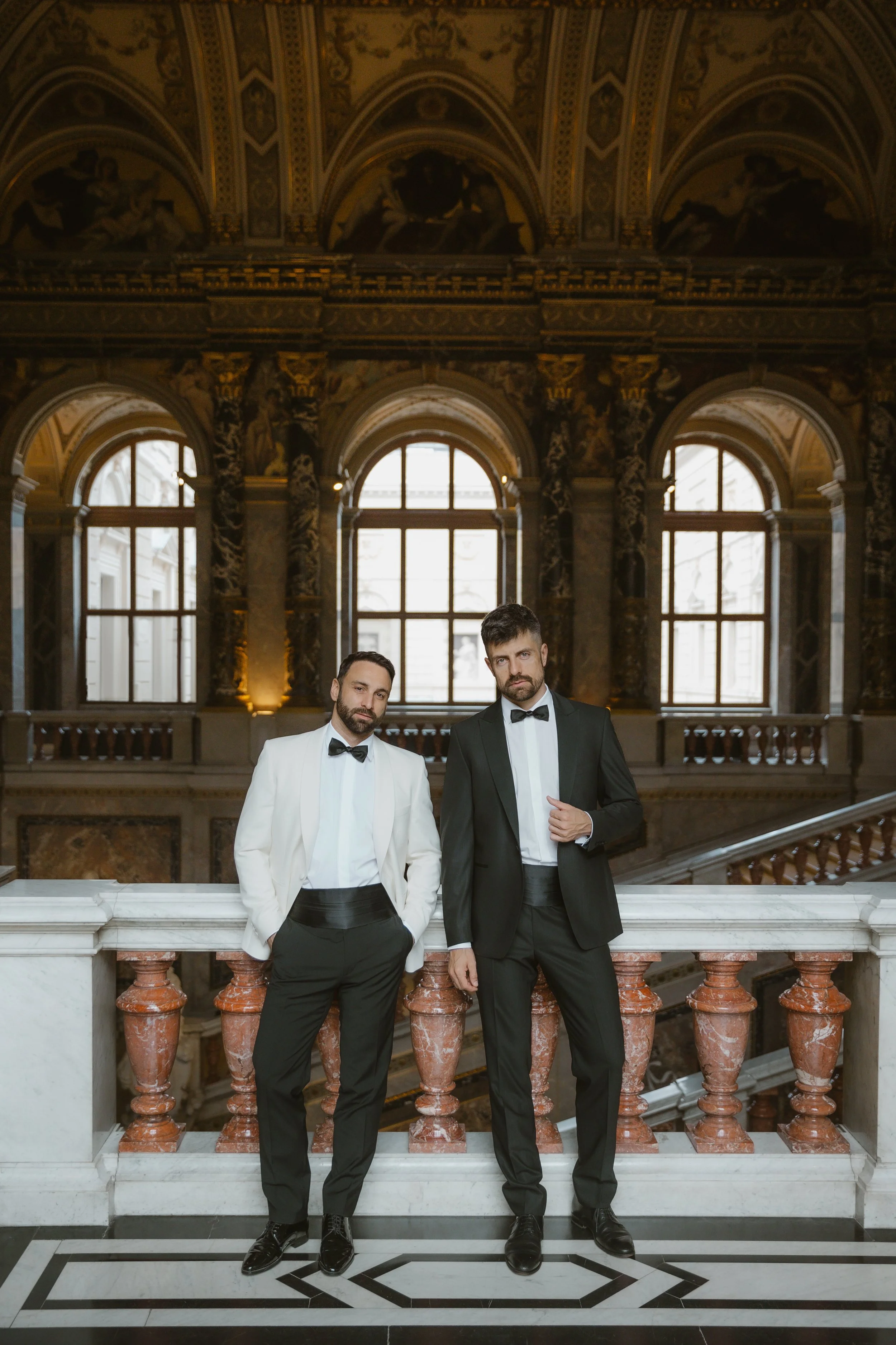 Zwei Männer in formeller Abendgarderobe stehen auf einer Marmortreppe in einem prunkvollen, historischen Saal mit hohen Fenstern und kunstvollen Wand- und Deckenmalereien.