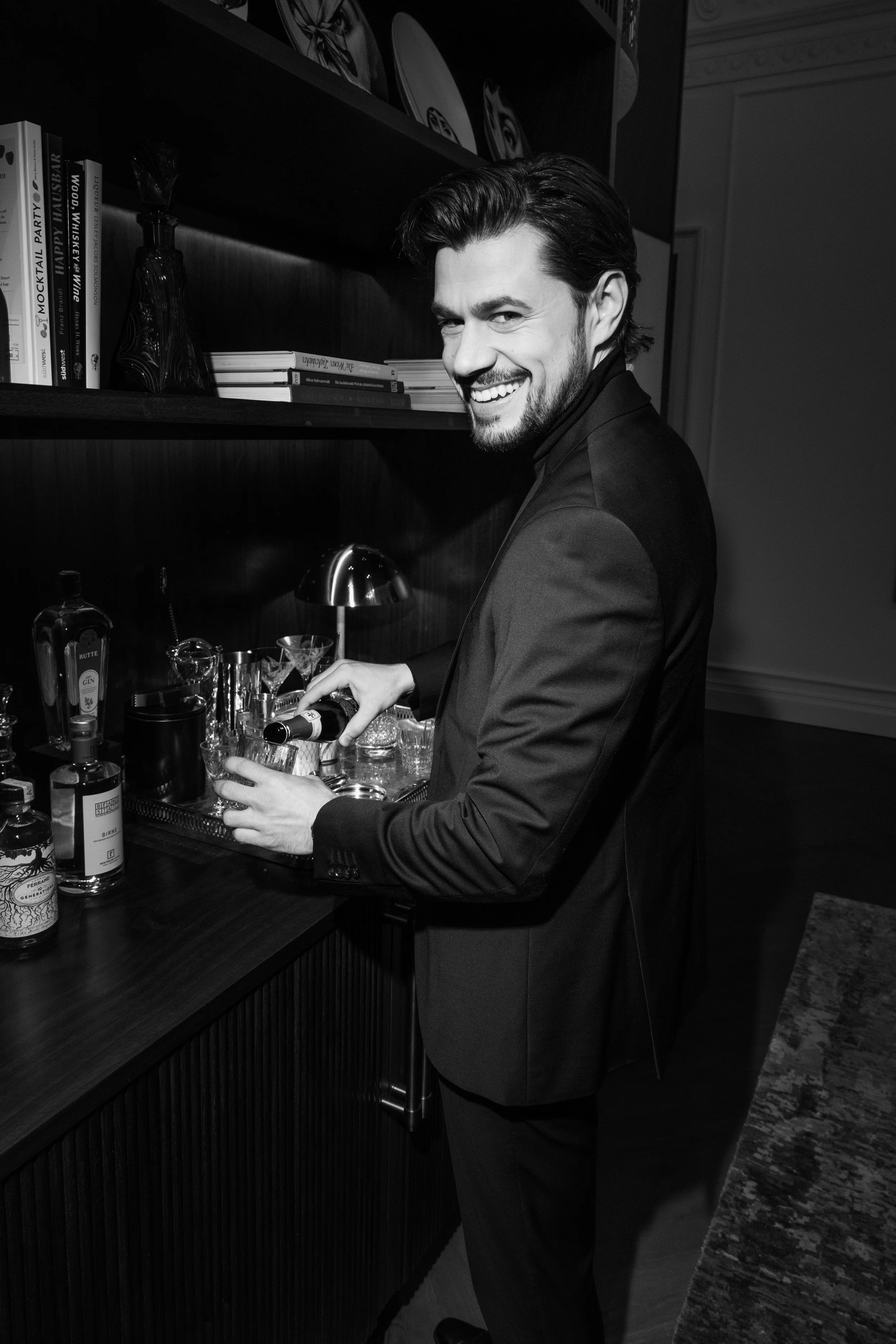 Ein Mann in einem Anzug bereitet an einer Hausbar ein Getränk zu. Die Umgebung ist stilvoll eingerichtet mit Regalen voller Bücher, Flaschen und Gläsern.