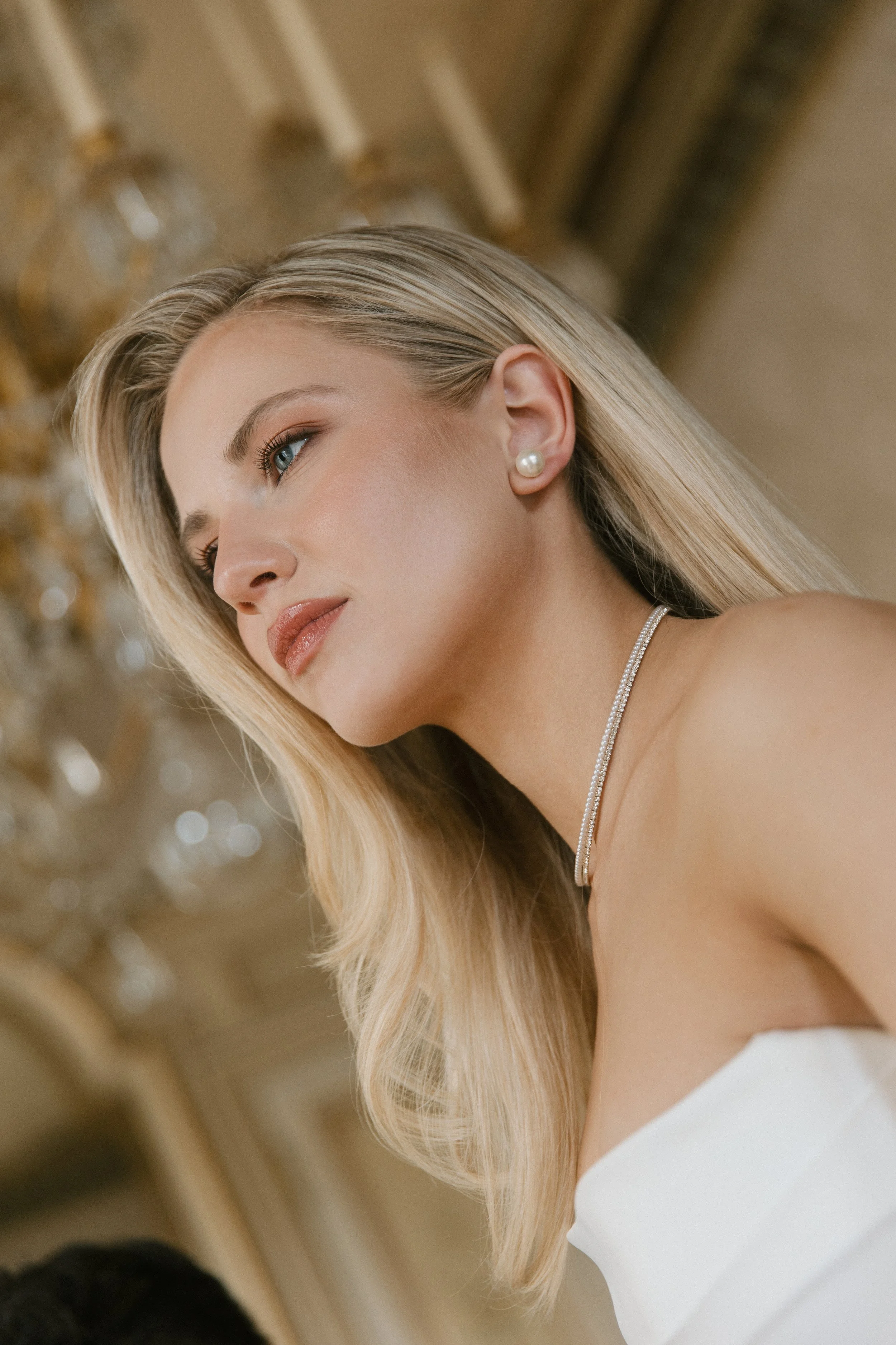 Eine junge blonde Frau mit blauer Augen, die elegantePerlenohrringe und eine Halskette trägt, sitzt in einem prunkvollen Raum mit goldener Decke und Kronleuchter.