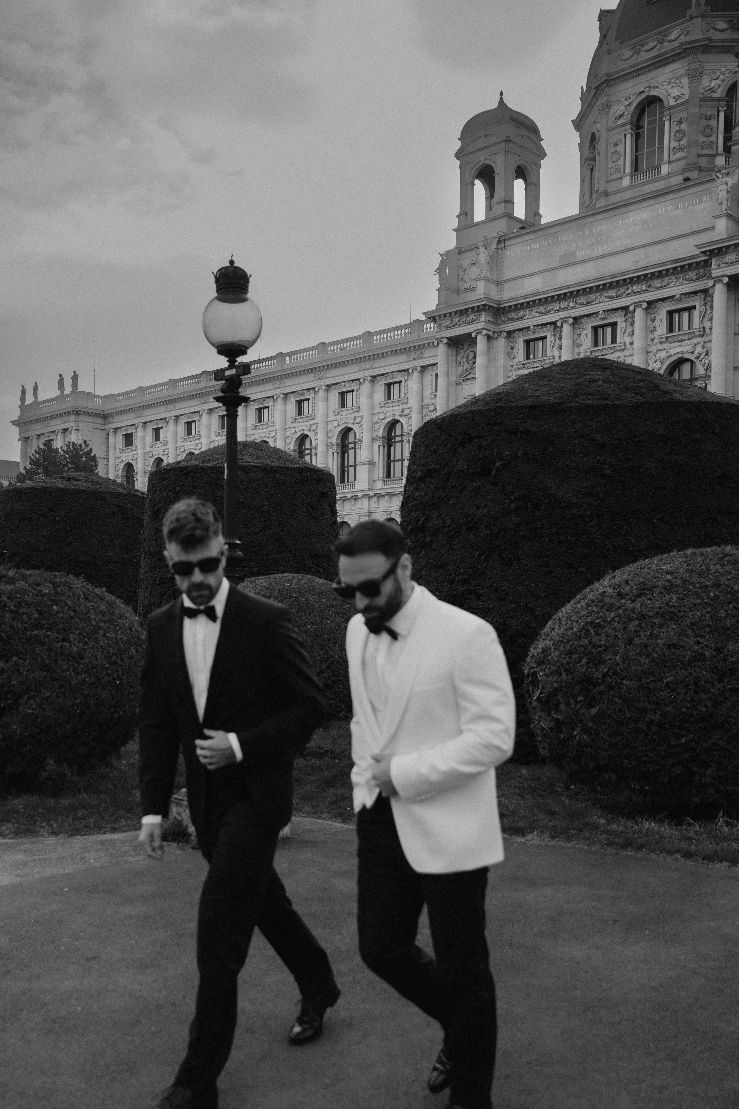 Zwei Männer in formeller Kleidung, einer in einem schwarzen Anzug und einer in einem weißen Anzug, laufen auf einem Weg vor einem historischen Gebäude mit Stuckarbeiten und großen Fenstern, umgeben von großen, runden Büschen. Es ist eine nächtliche Szene.