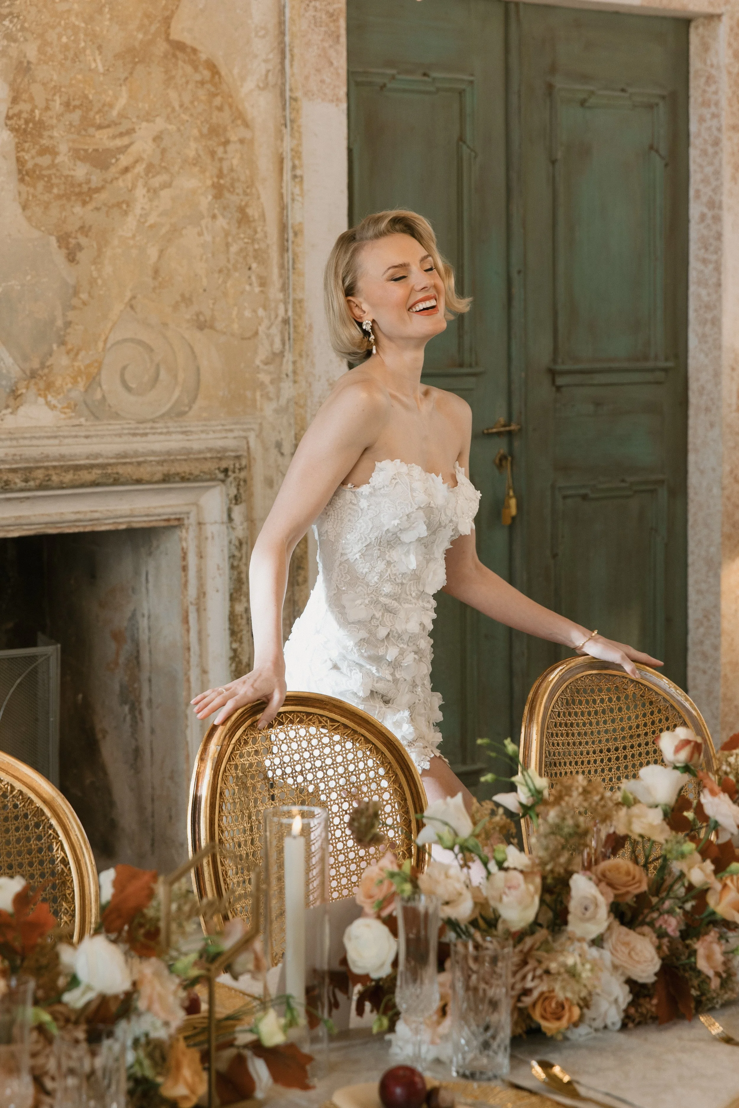Eine lachende Frau in einem weißen, mit Blumen verzierten Kleid, die an einem Tisch mit Blumen und Kerzen sitzt, in einem historischen Raum mit grünen Türen und dekorativen Wänden.