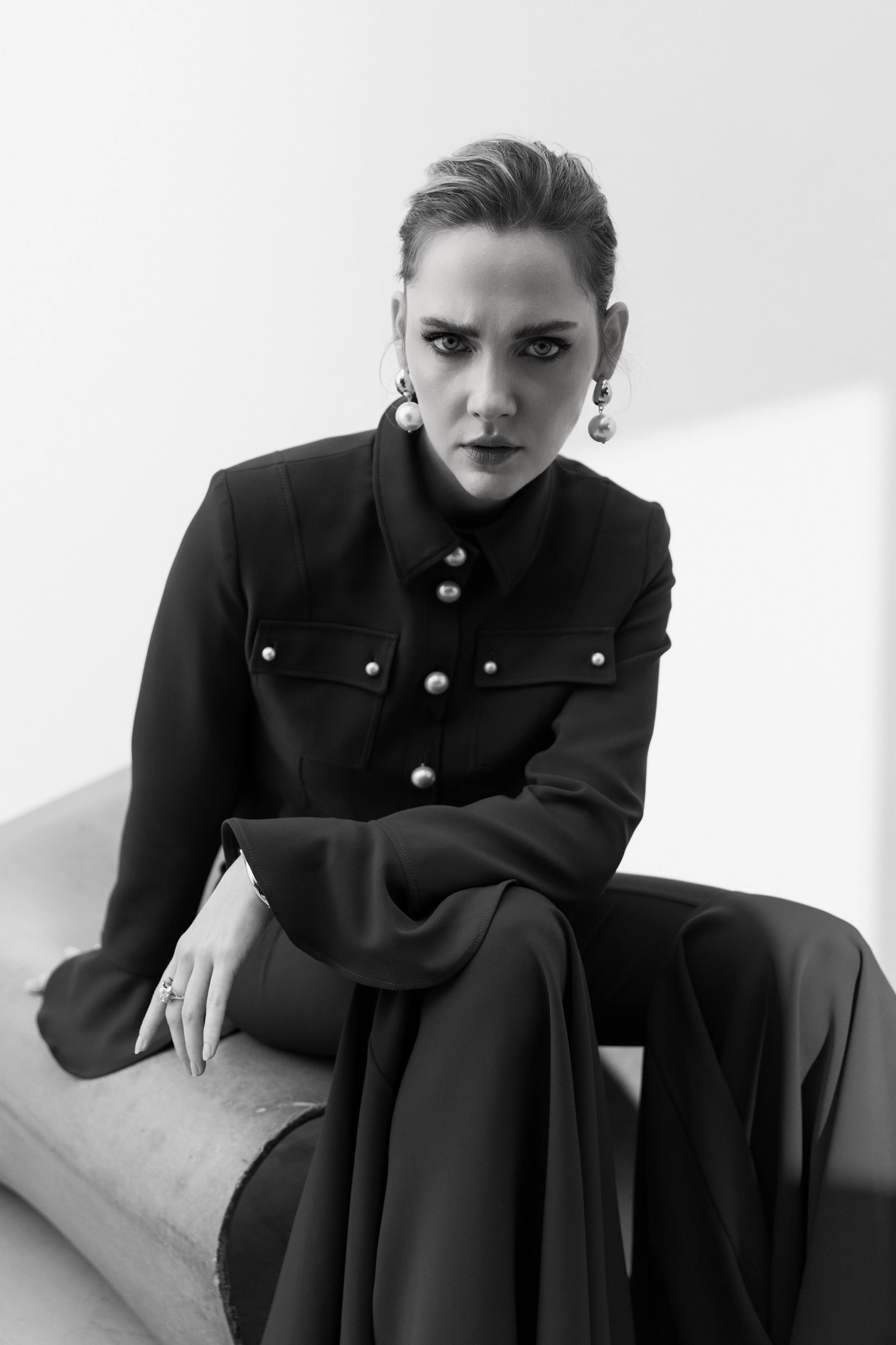 Schwarz-Weiß-Foto einer Frau mit dicker Augenbraue, hochgestecktem Haar, großen Perlenohrringen, schwarzer Jacke mit Perlendetails, sitzt auf einem langen Sitz, blicke direkt in die Kamera, skeptischer Ausdruck.