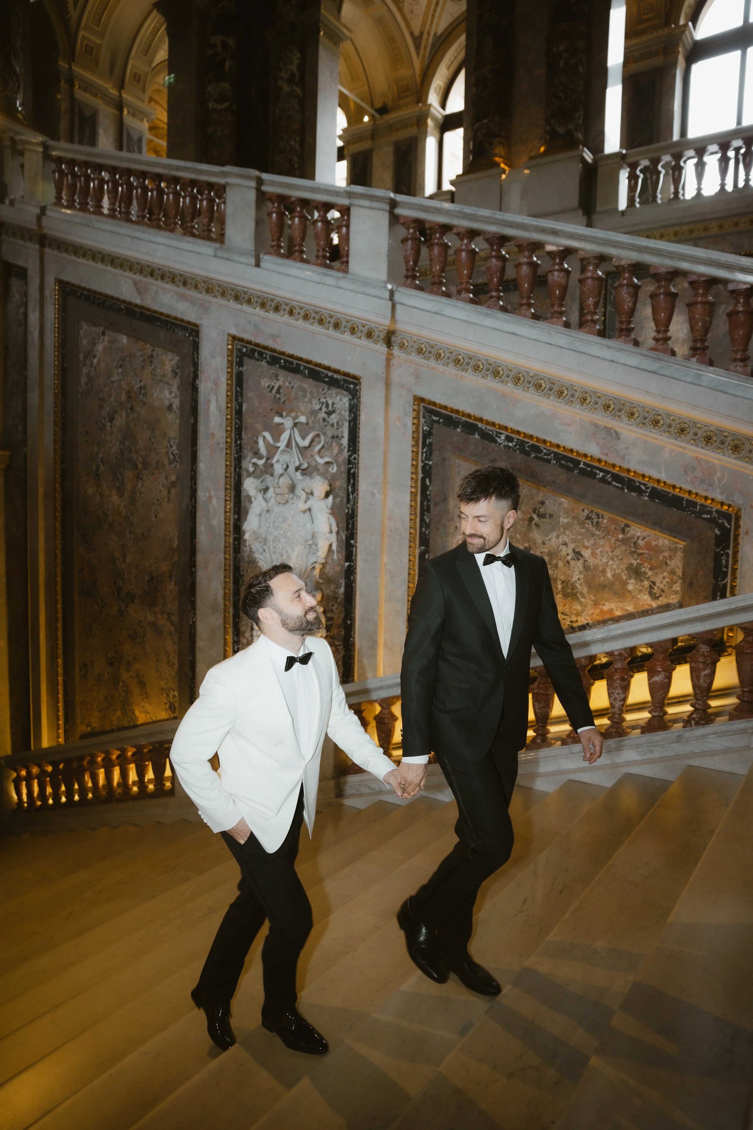 Zwei Männer in formeller Kleidung, einer in weißem Anzug, der andere in schwarzem Anzug, halten Händchen beim Treppensteigen in einem eleganten, historisch dekorierten Innenraum.